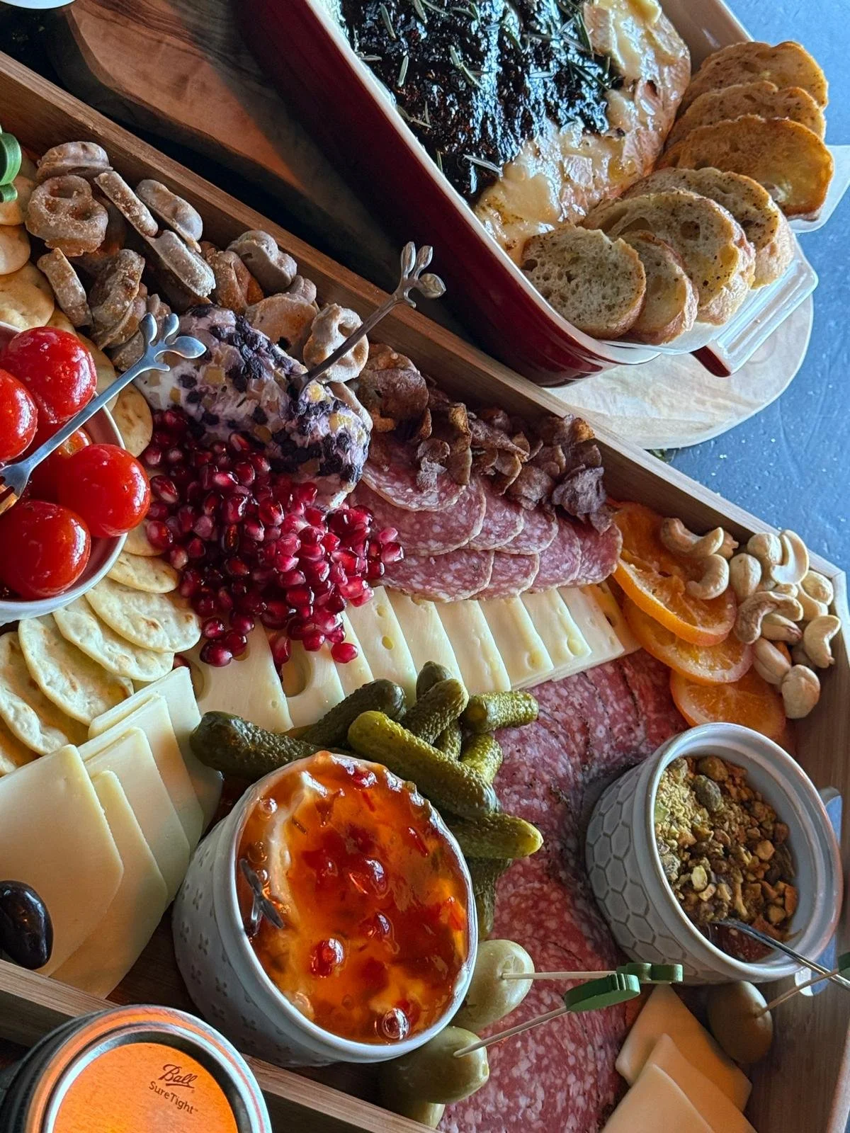 A cheese and charcuterie platter with fresh tomatoes, pickles, nuts, dried and fresh fruits, cured meats, cheeses, hummus, and a baked brie with jam, served on a wooden board.