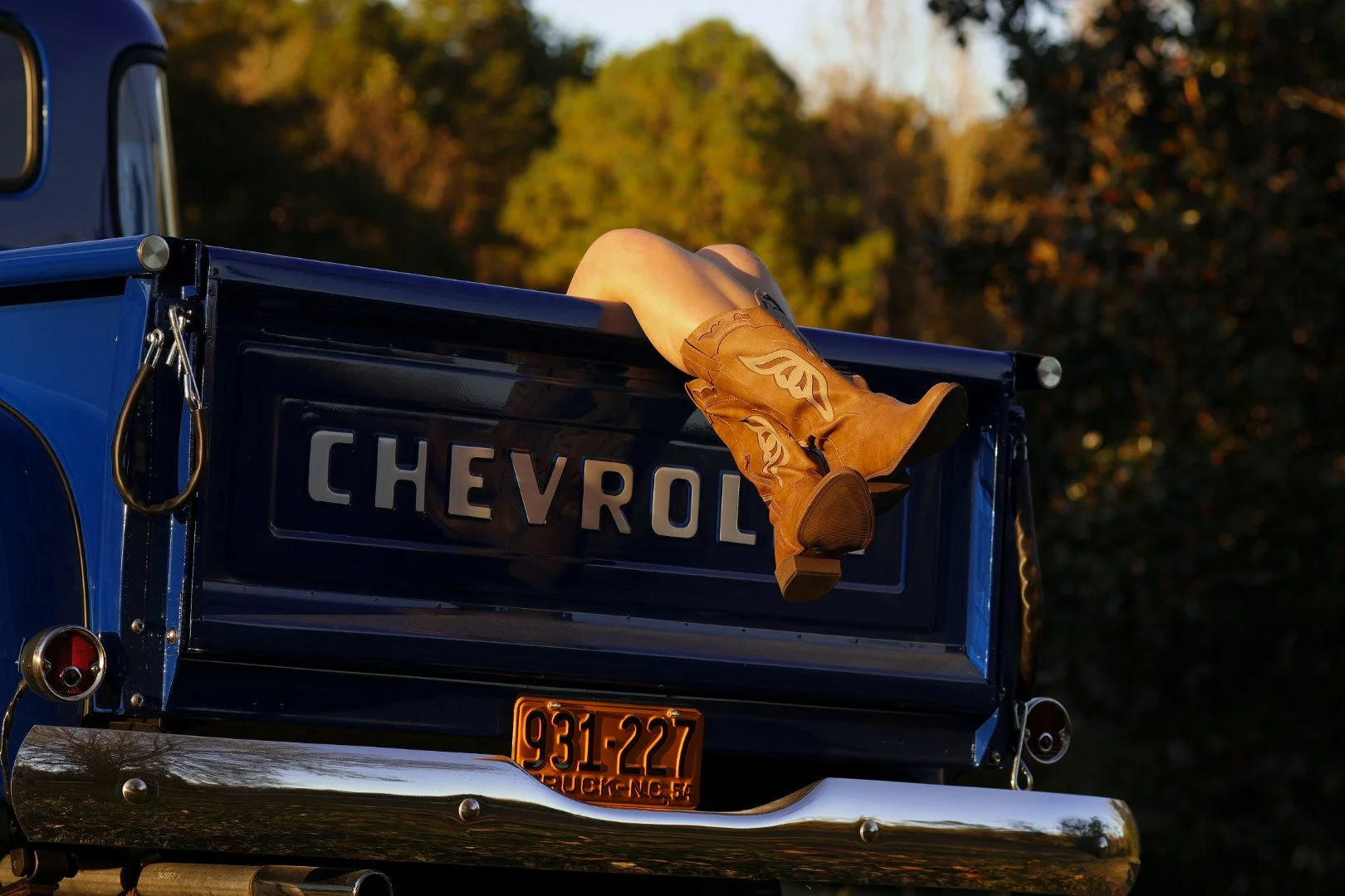 A vintage blue Chevy pickup truck with a pair of tan cowboy boots hanging from the tailgate, set against a background of trees in autumn.