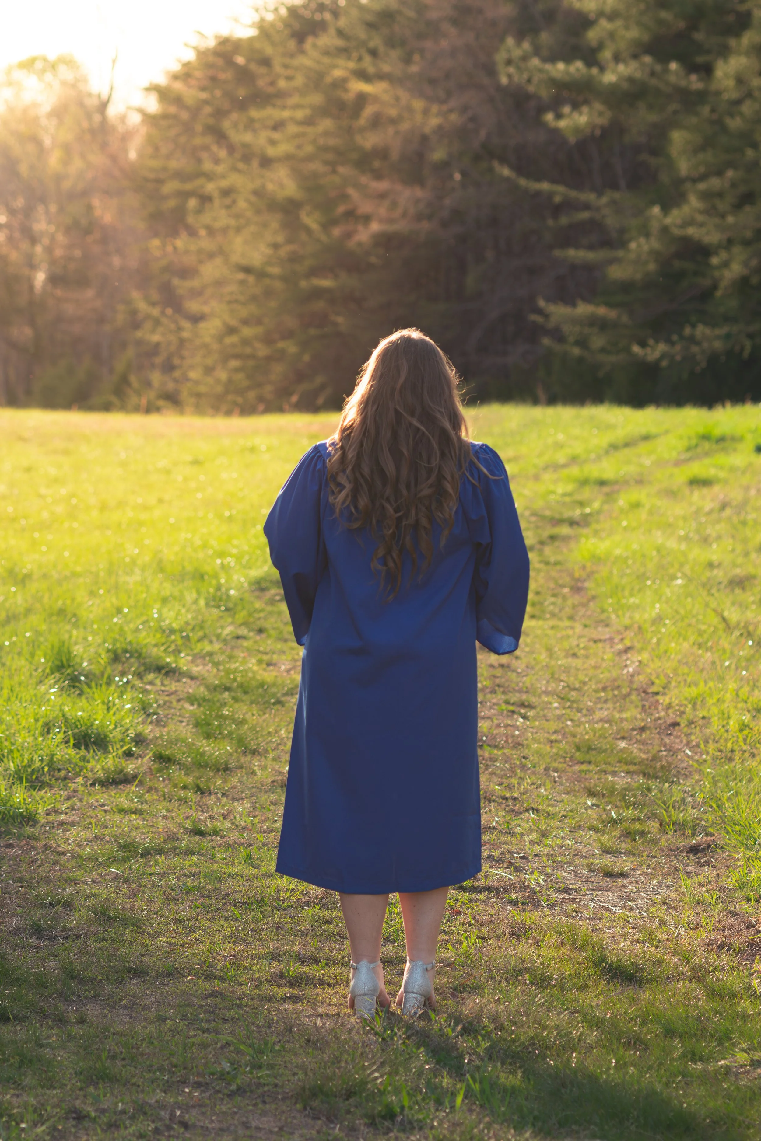 A woman in a blue dress and heels walking on a grassy path in a park during sunset or sunrise, with tall trees and sunlight in the background.