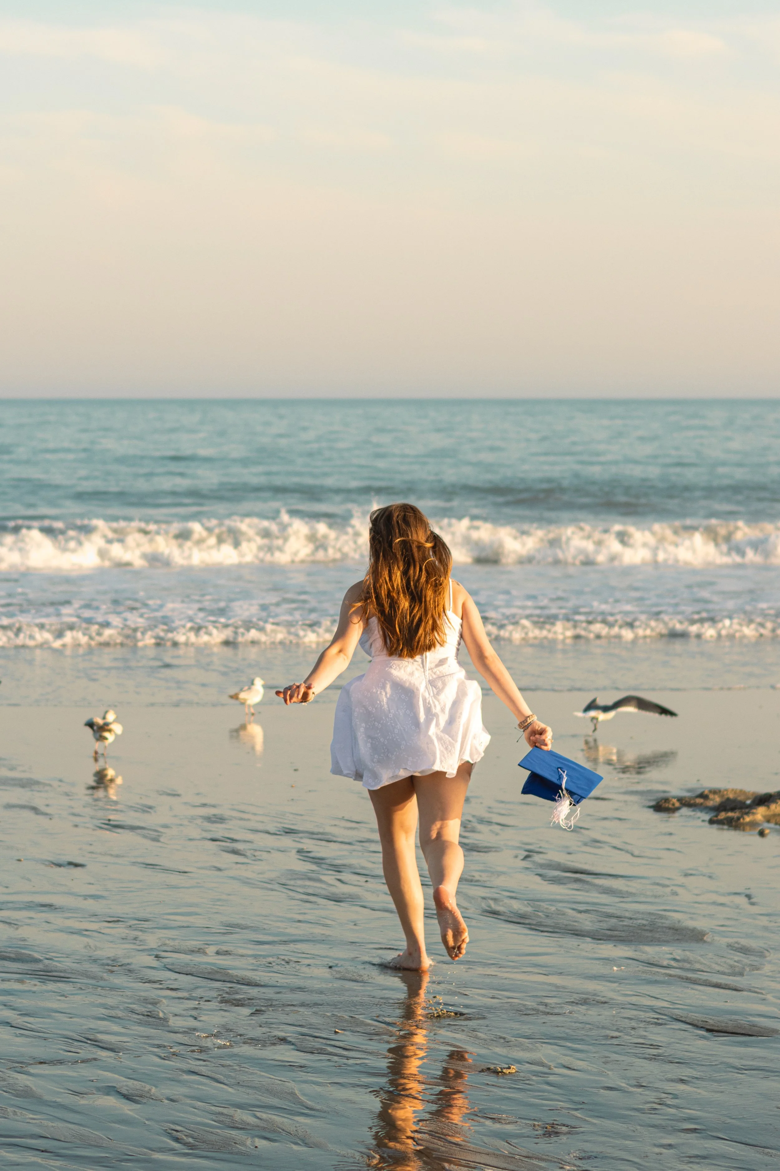 Woman in a white dress walking in shallow ocean water, holding a blue graduation cap with a tassel, surrounded by seagulls, ocean waves, and a cloudy sky.