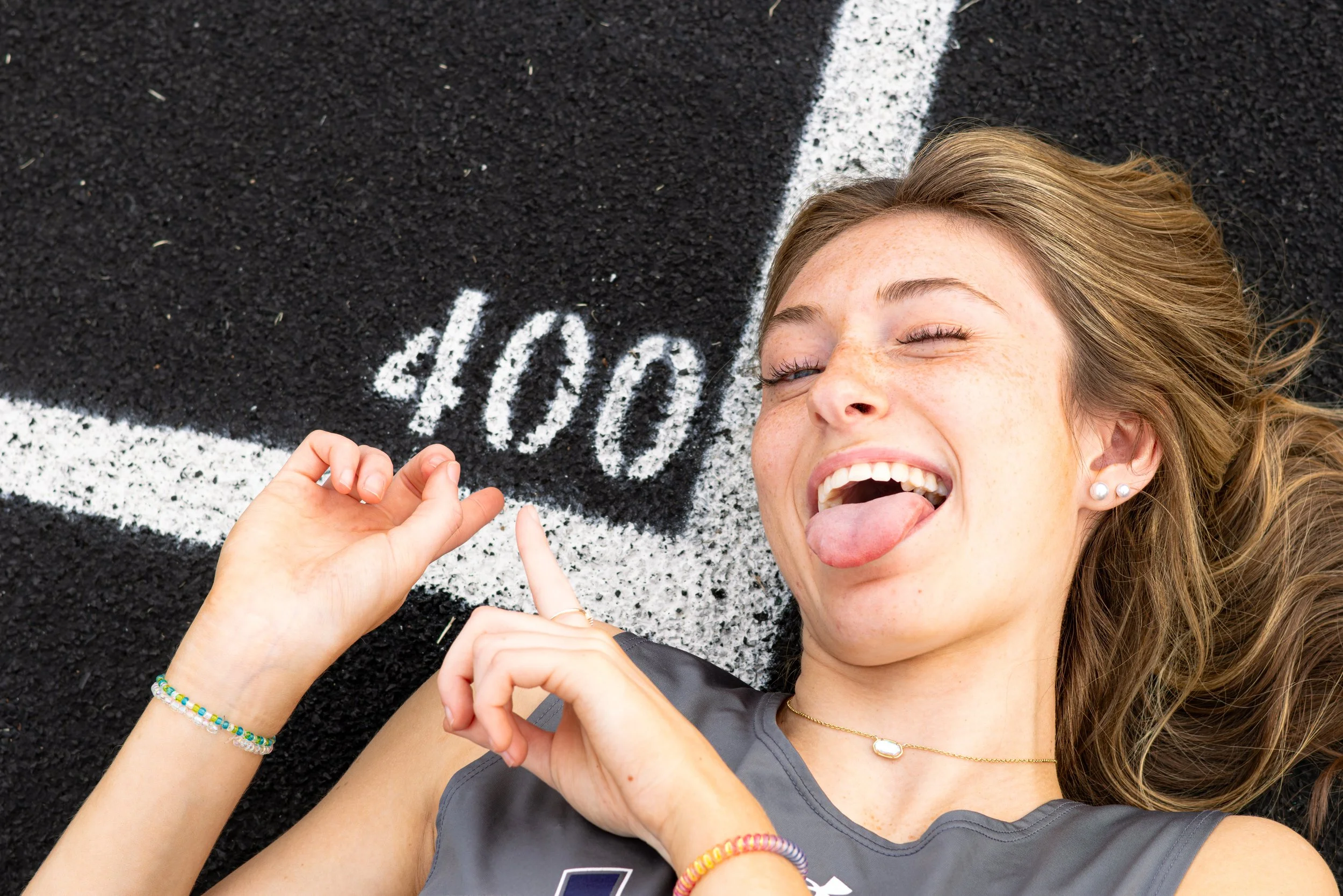 Young woman lying on a running track making a heart shape with her hands, sticking her tongue out, with the word 'love' written in white chalk on the black surface.