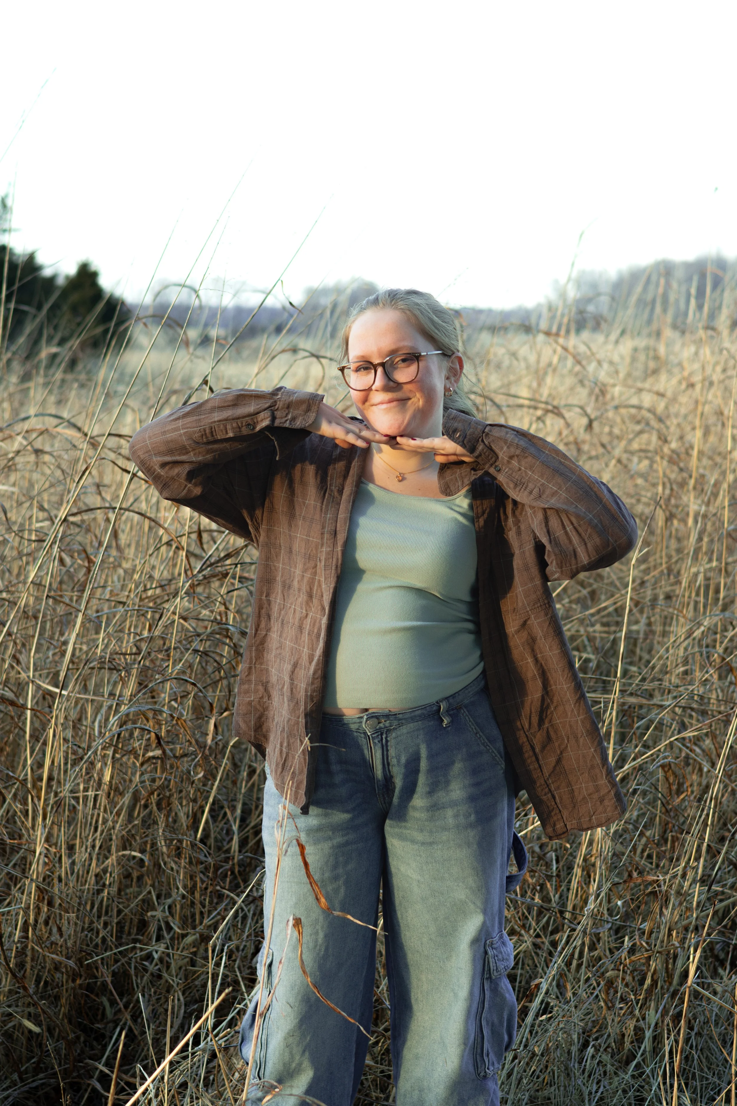 A woman with glasses in a brown plaid shirt and ripped jeans standing in a field of tall, dry grass, smiling with her hands under her chin, in a natural outdoor setting.