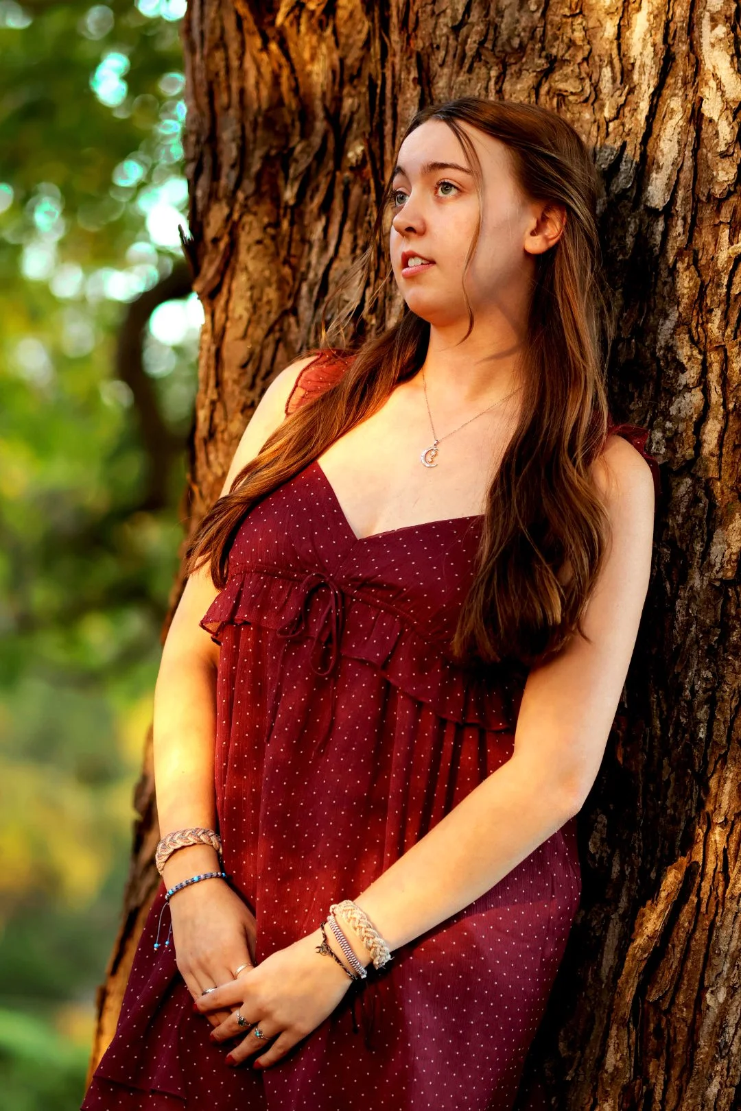 A young woman with long brown hair leaning against a large tree trunk in a forest. She is wearing a maroon, polka-dotted dress and is looking to her left. She has a necklace with a crescent moon pendant and several bracelets on her wrists.