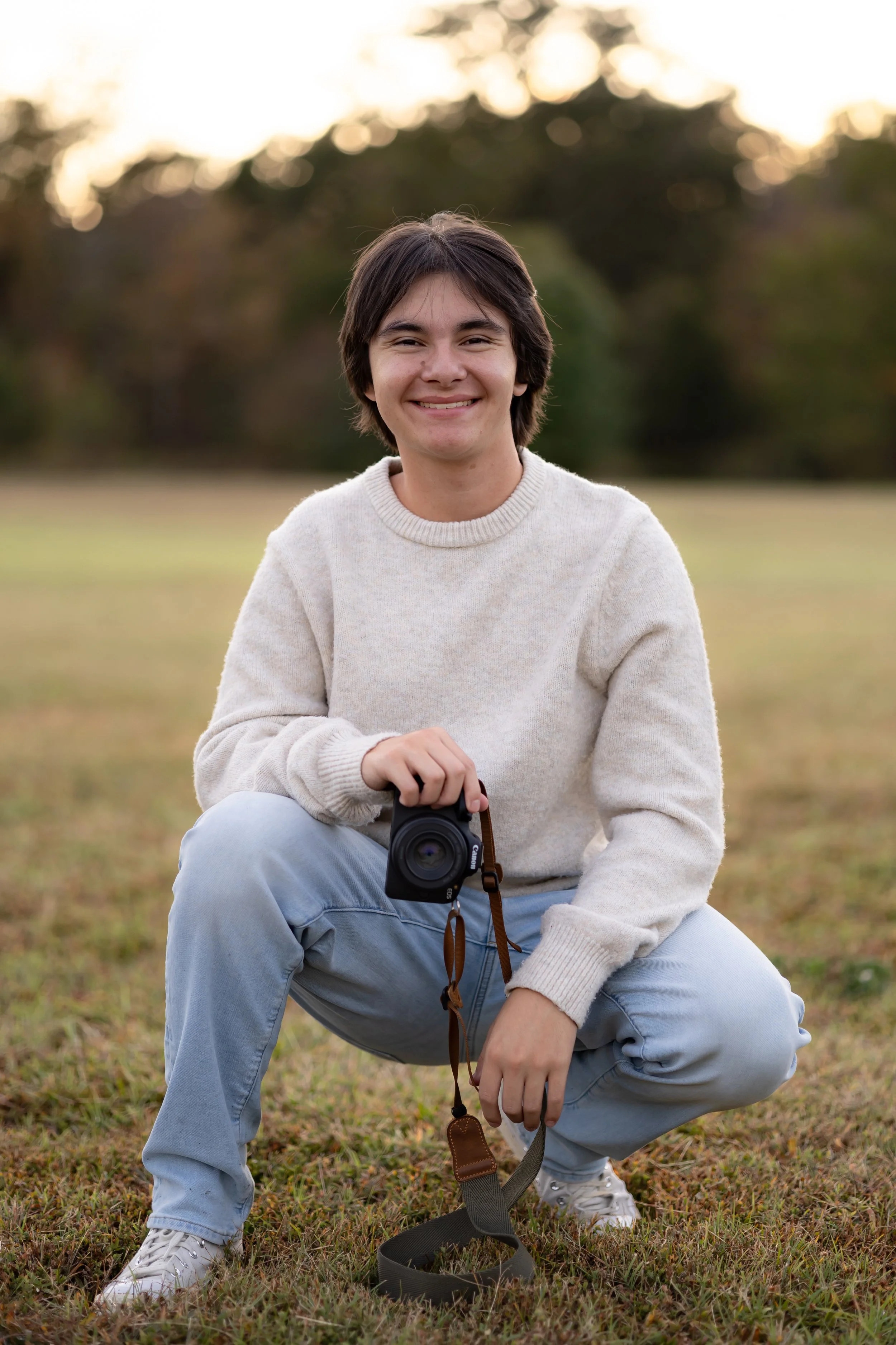 Young man with dark hair and a smile, wearing a cream sweater, light blue jeans, and white sneakers, squatting outdoors on grass, holding a black camera with a brown strap, with a blurred background of trees and a sunset sky.