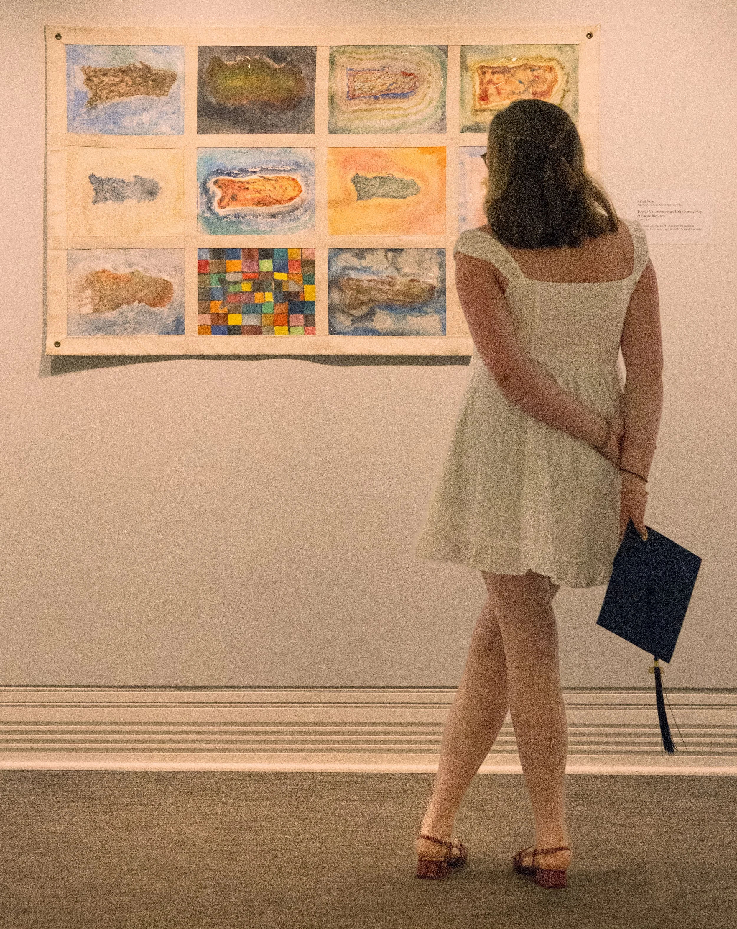 A woman wearing a white dress and sandals viewing a colorful art display on a wall inside an art gallery.