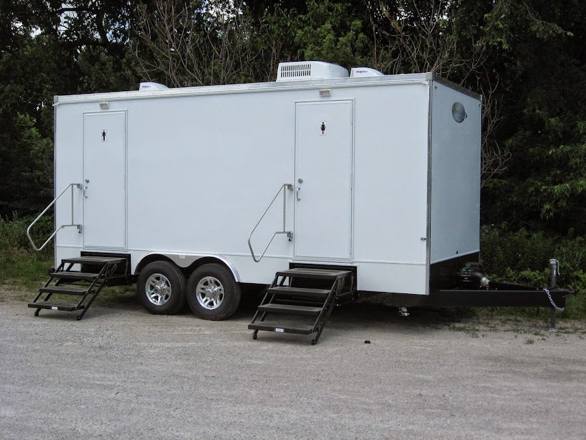 Empty white mobile food or restroom trailer with two doors, stairs, and a small air conditioning unit, parked on a dirt surface with trees in the background.
