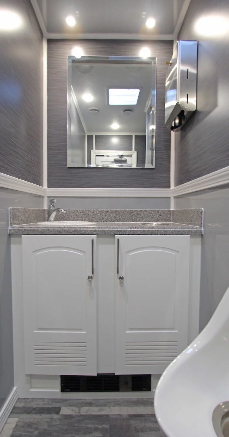 A bathroom with a white vanity with a granite countertop, a sink, a mirror, a wall-mounted soap dispenser, and a toilet on the right. The walls have gray and white paneling, and the ceiling has recessed lighting.