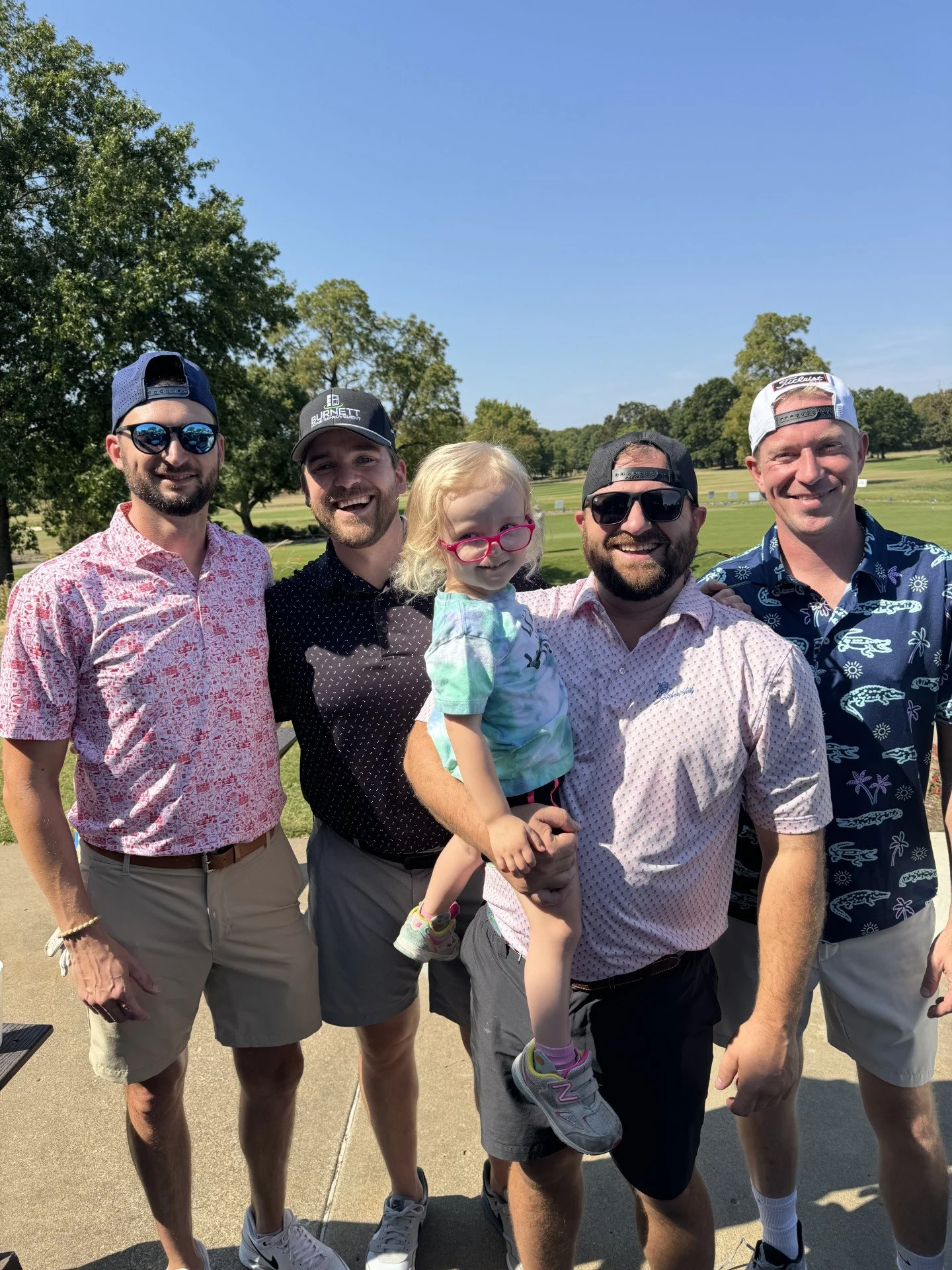 Group of five men and a young girl smiling outdoors, dressed in casual summer clothes, at a park or golf course with trees and grass in the background.