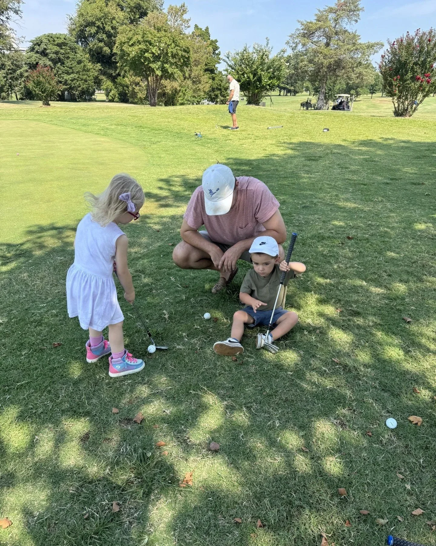A man and two young children on a golf course, preparing to hit golf balls. The man is crouching next to a young boy, and a girl watches as she holds a golf club. The scene is set on a grassy area with trees and a golf cart in the background.