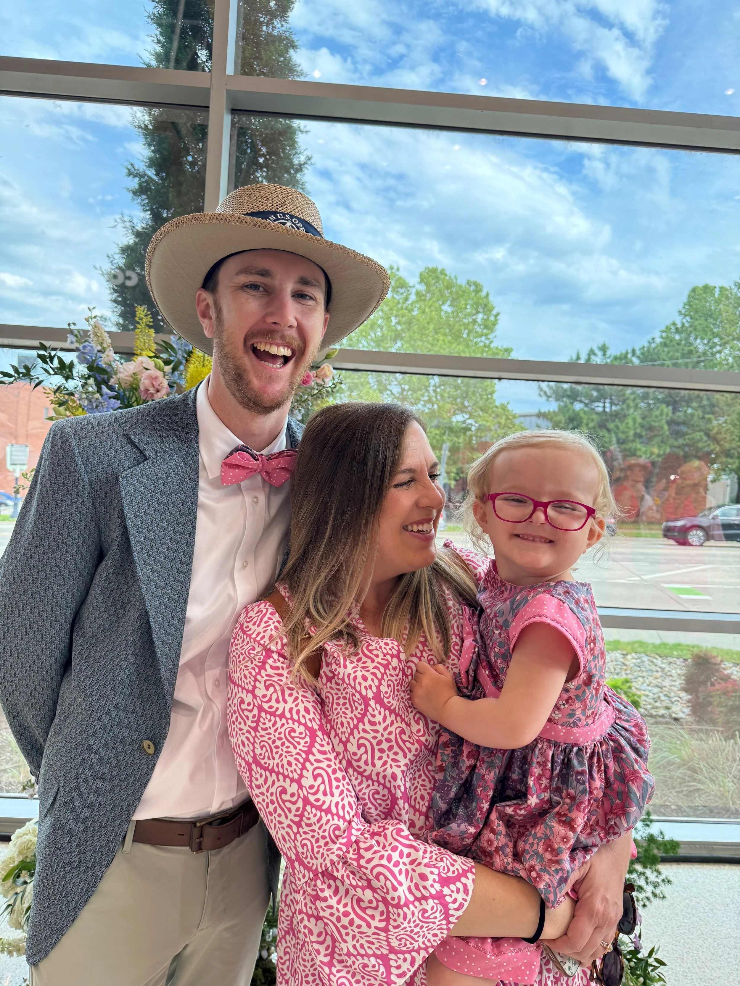 A group of four people, including a man in a gray blazer and pink bow tie, a woman with long brown hair, a young girl with glasses, and part of another woman, are smiling and posing inside a building with large windows showing trees and a cloudy sky outside.