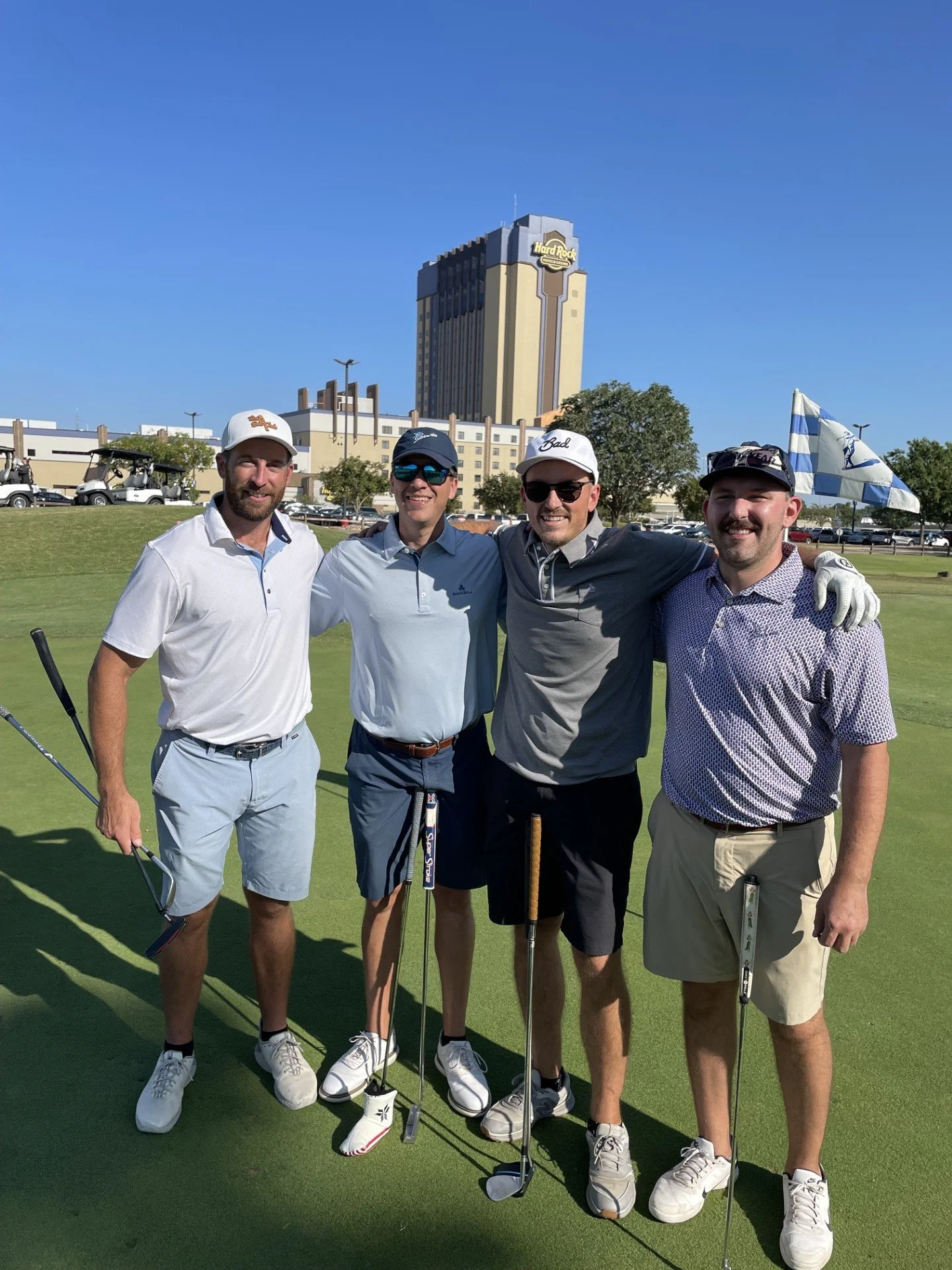 Four men standing on a golf course, smiling, with golf clubs in hand. Behind them, a building with a "Hard Rock" sign and a clear blue sky.