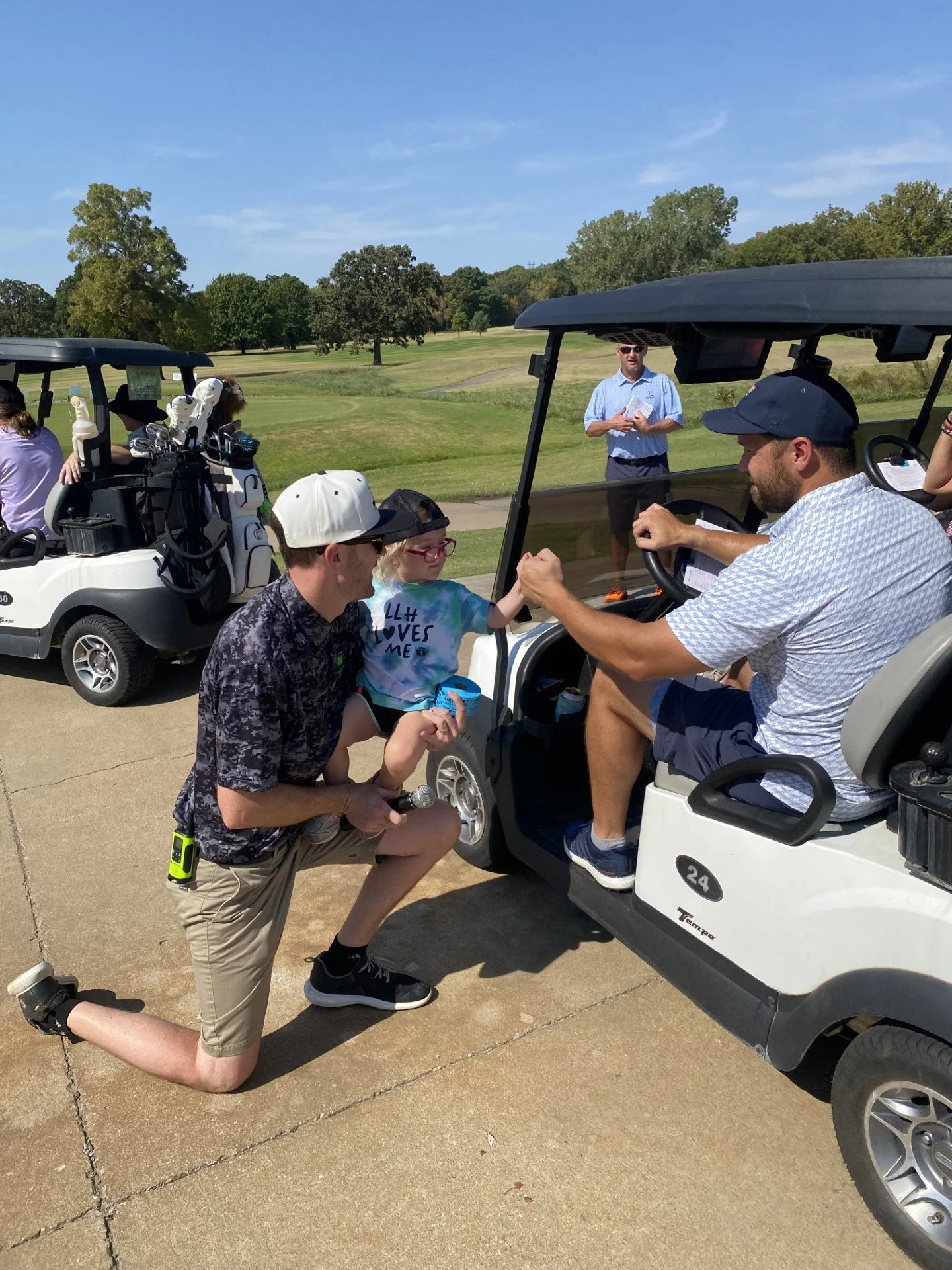 A man kneeling on one knee handing a trophy to a young girl sitting in a golf cart. A woman is sitting in a golf cart behind them. Other people and golf carts are in the background on a golf course with trees and a blue sky.