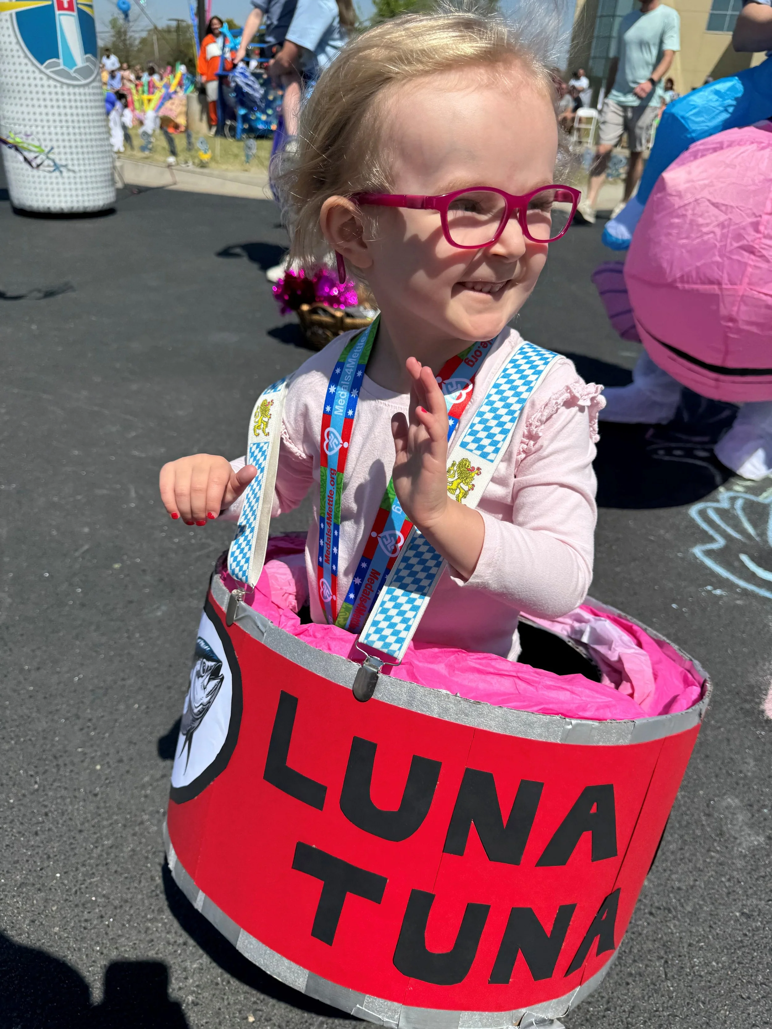 A young girl with blonde hair, pink glasses, and a big smile, sitting in a red UNICEF donation drum at an outdoor event. She is wearing a pink shirt with ruffled sleeves and has a UNICEF lanyard around her neck. Behind her, there are people and colorful decorations at a lively gathering.