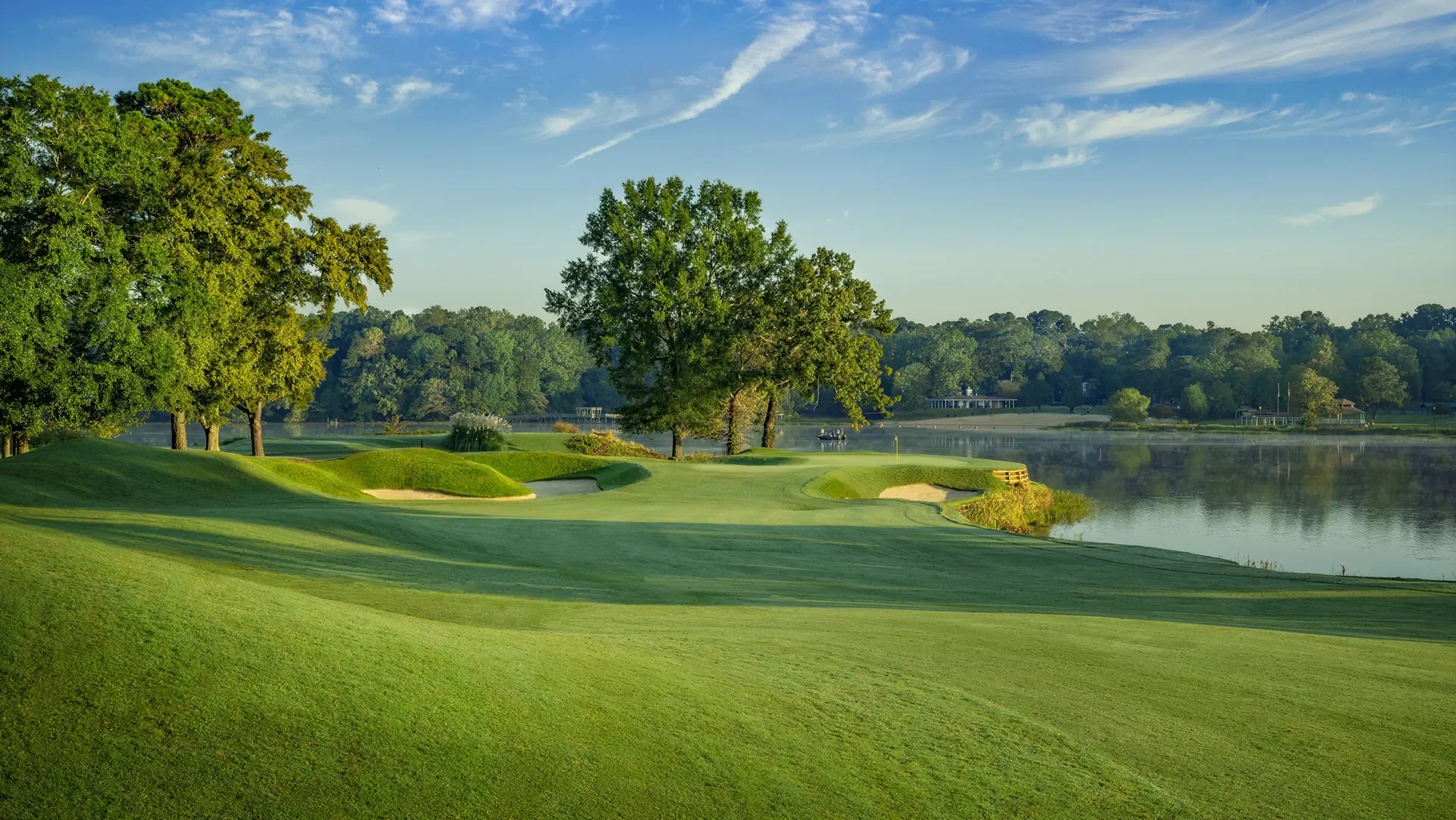 A scenic golf course by a lake with lush green grass, sand bunkers, and tall trees, under a blue sky with thin clouds.