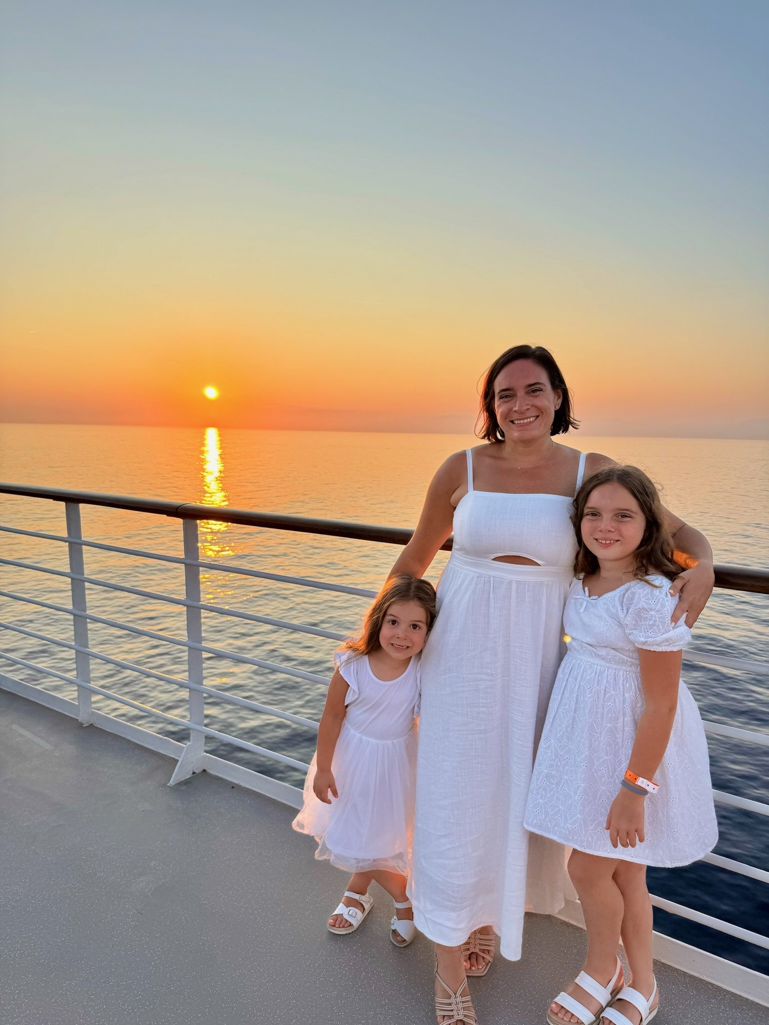 A woman and two young girls in white dresses standing on the deck of a boat during sunset.