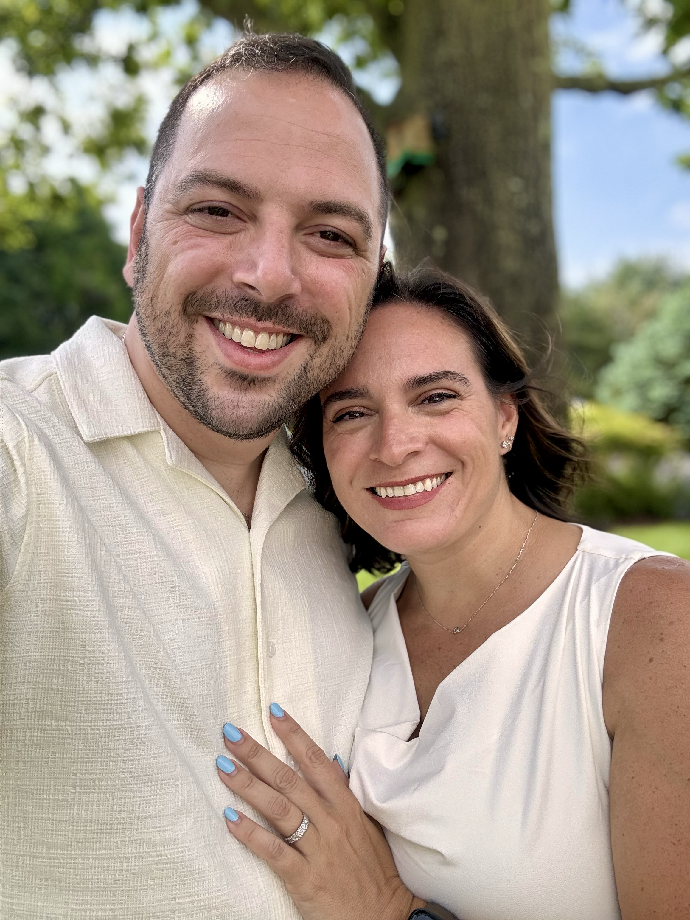 A smiling couple taking a selfie outdoors with a large tree in the background. The woman has dark hair, is wearing a sleeveless white top, and a ring on her finger. The man has short dark hair and a beard, and is wearing a light-colored collared shirt.