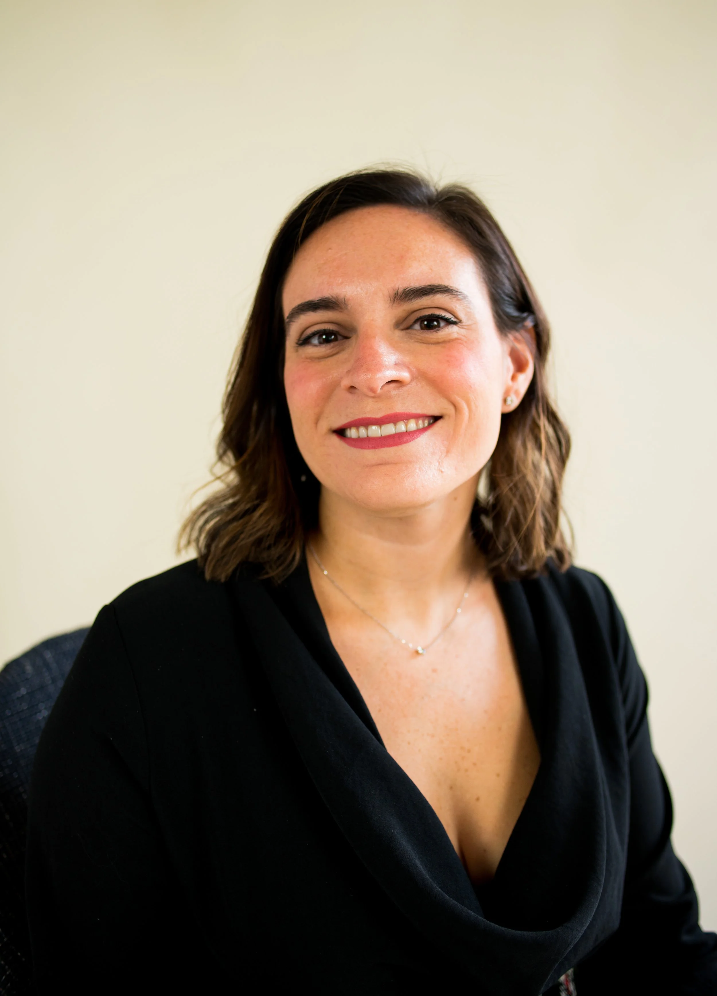A woman with shoulder-length brown hair, smiling, wearing a black top and a silver necklace, sitting against a plain light-colored background.