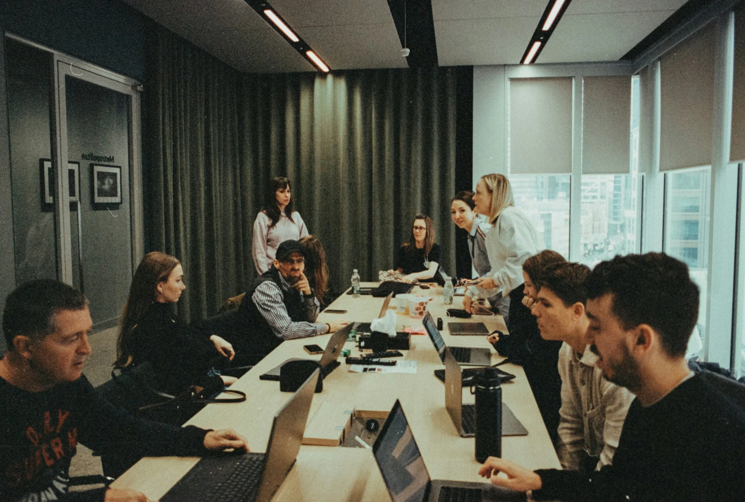 People gathered in a modern conference room for a meeting or workshop, with some working on laptops and others listening or speaking.