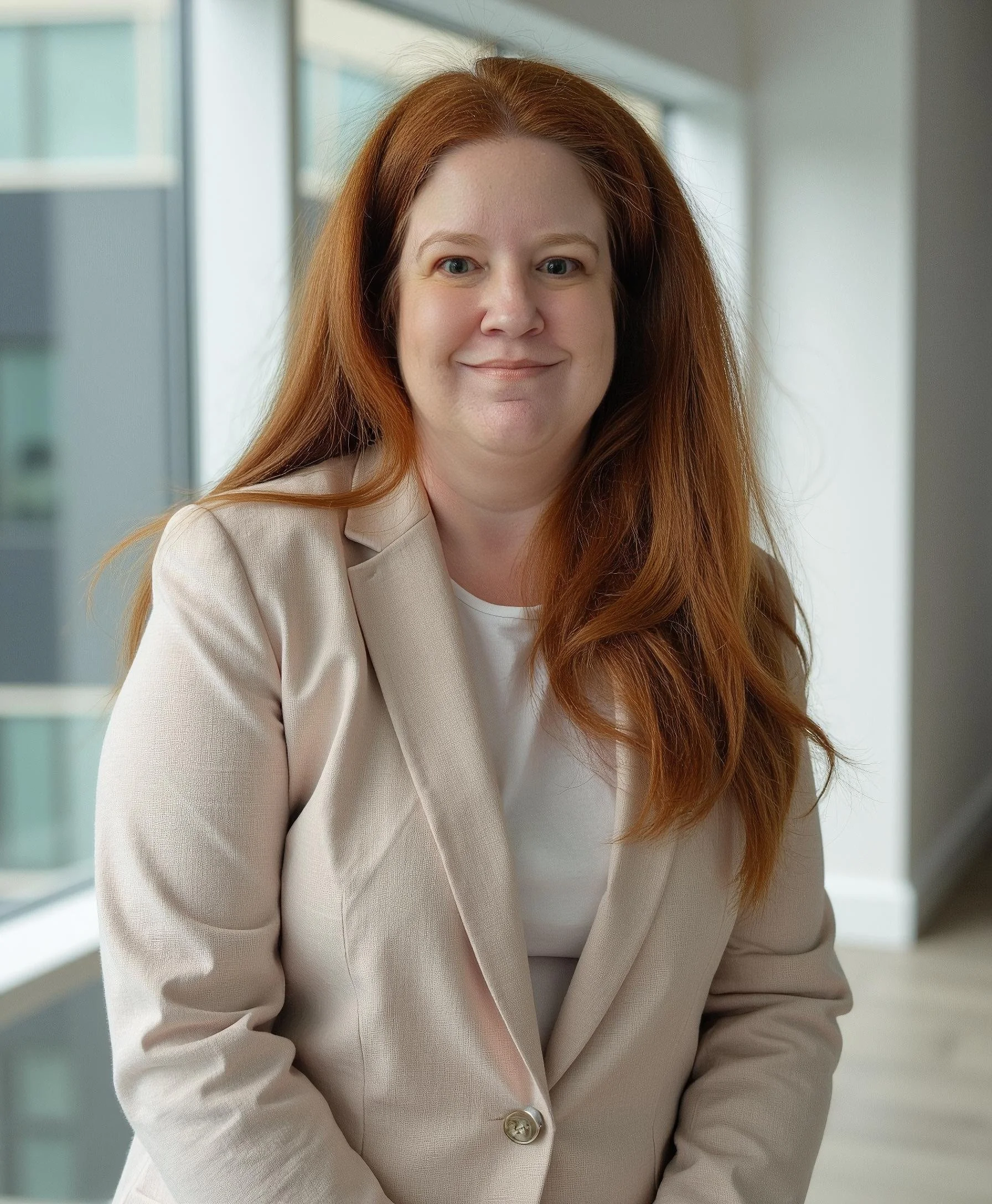 A portrait of a woman with long reddish-brown hair, wearing a beige blazer, smiling softly, in front of a modern interior with large windows.