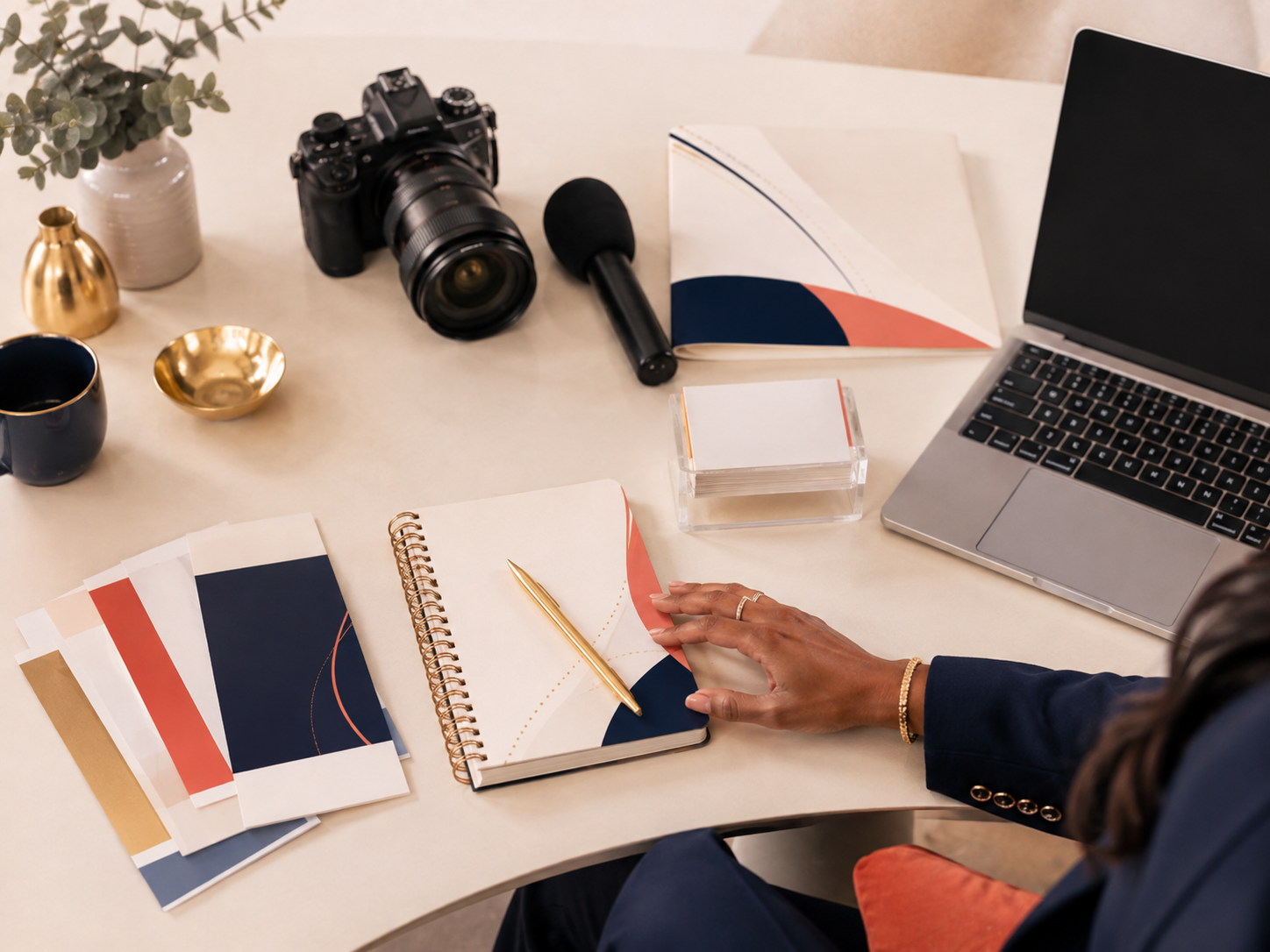 A workspace with a laptop, camera, microphone, notebooks, color swatches, a cup of coffee, and decorative items on a white table, with a person's hand reaching for a notebook.