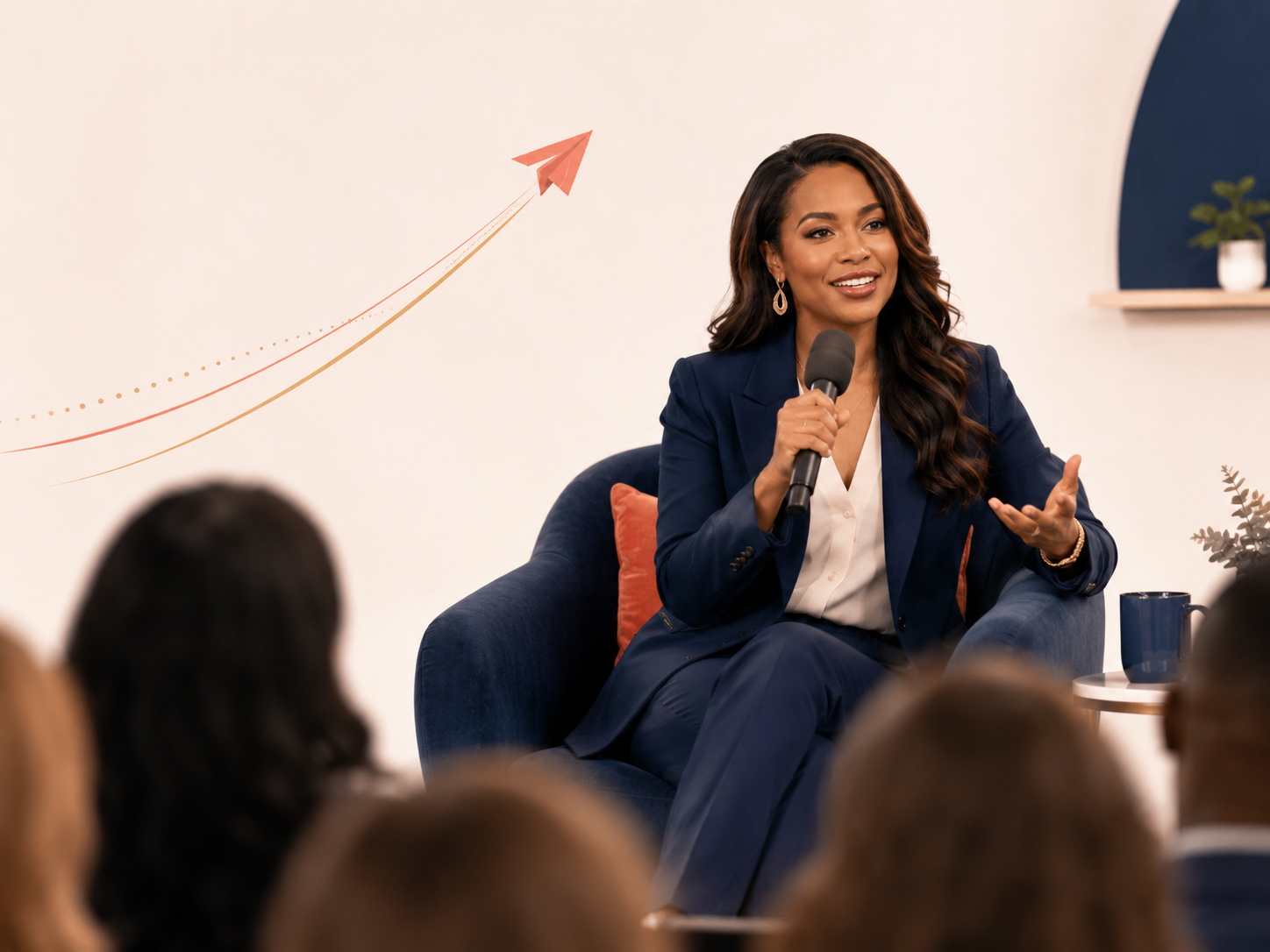 A woman in a navy blazer and white blouse is speaking into a microphone while seated in a blue armchair during a presentation. There is a graph on the wall showing an upward trend represented by an arrow.