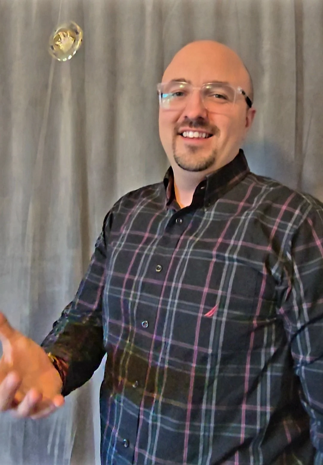 A man with glasses and a goatee, wearing a black and pink plaid shirt, smiling and shaking hands, standing in front of a gray curtain with two gold balloons.