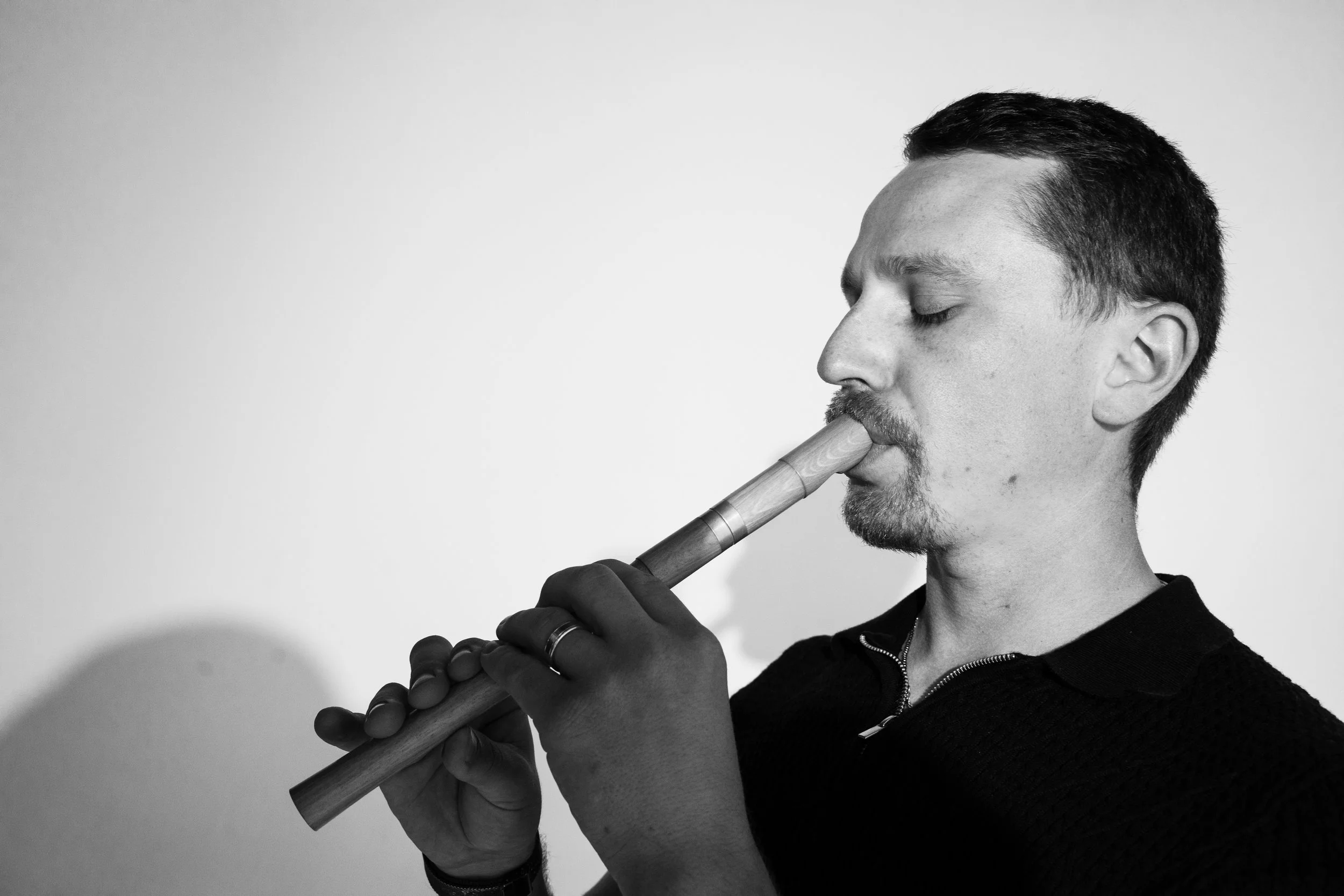A black and white photo of a man with short dark hair and facial hair, eyes closed, playing a wooden wind instrument against a plain background.