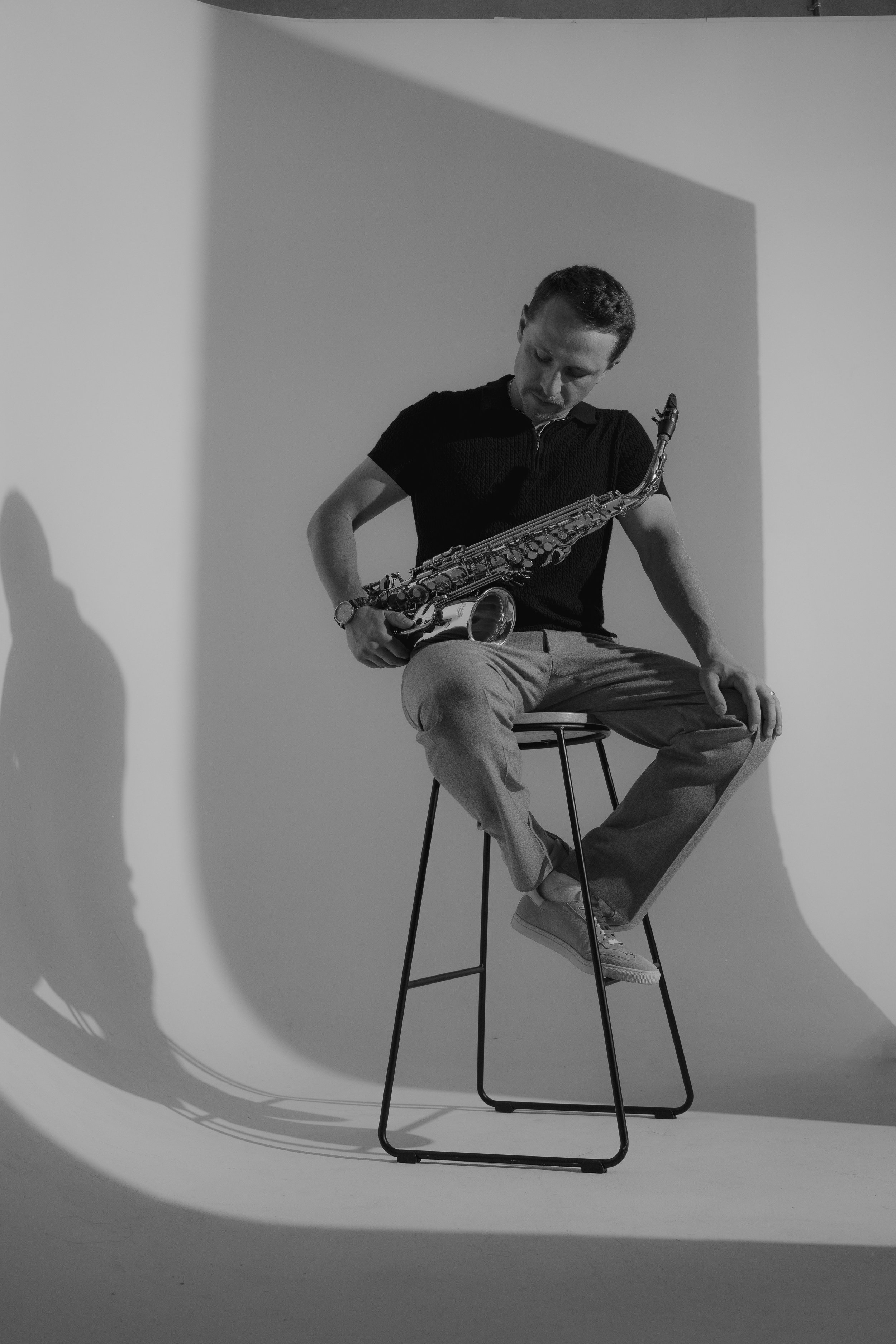 A young man sitting on a stool holding a saxophone, with a plain background and shadow cast on the wall.