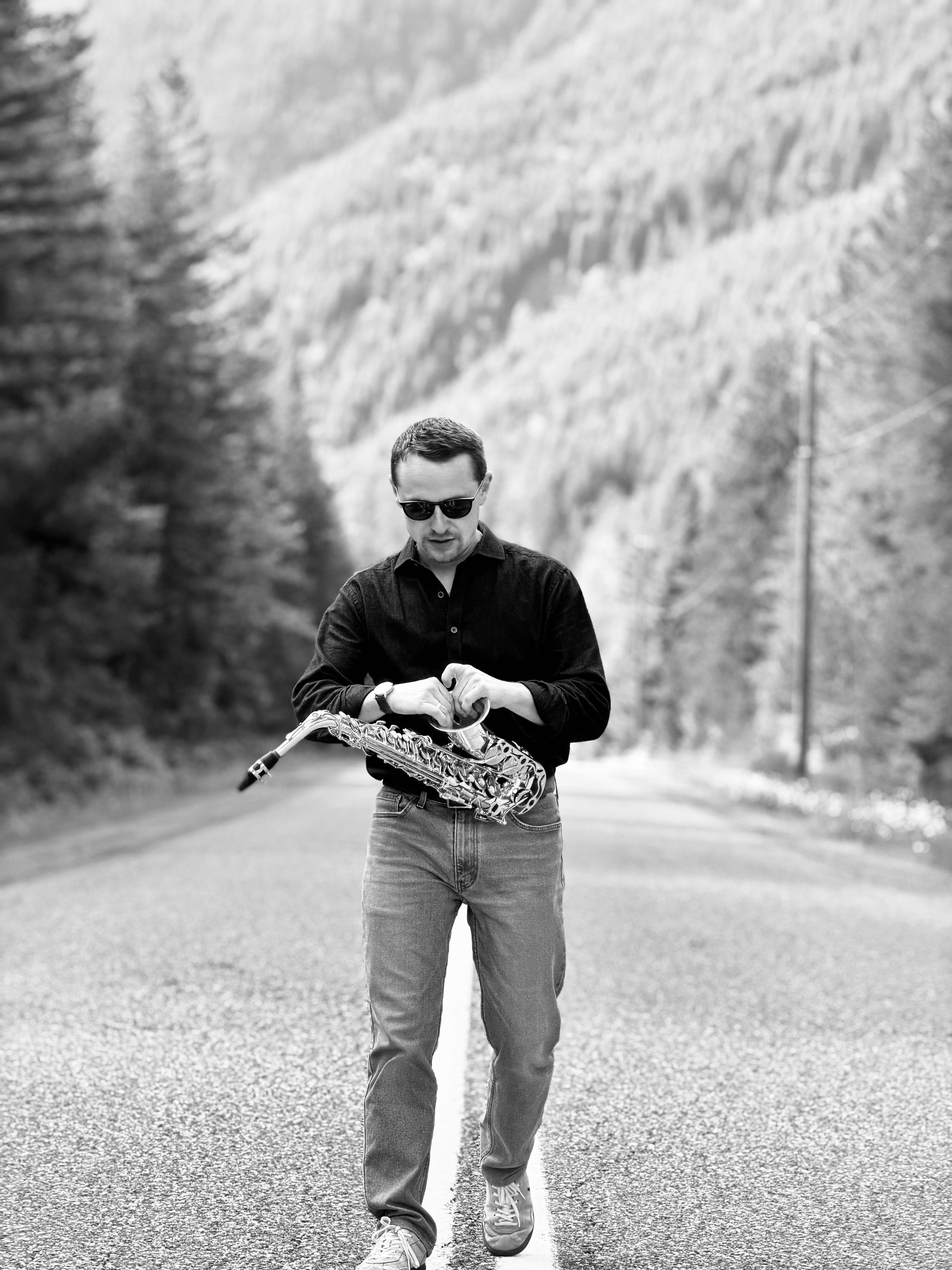 A man wearing sunglasses, a dark shirt, jeans, and sneakers walking down a deserted road while holding a saxophone in his hands.