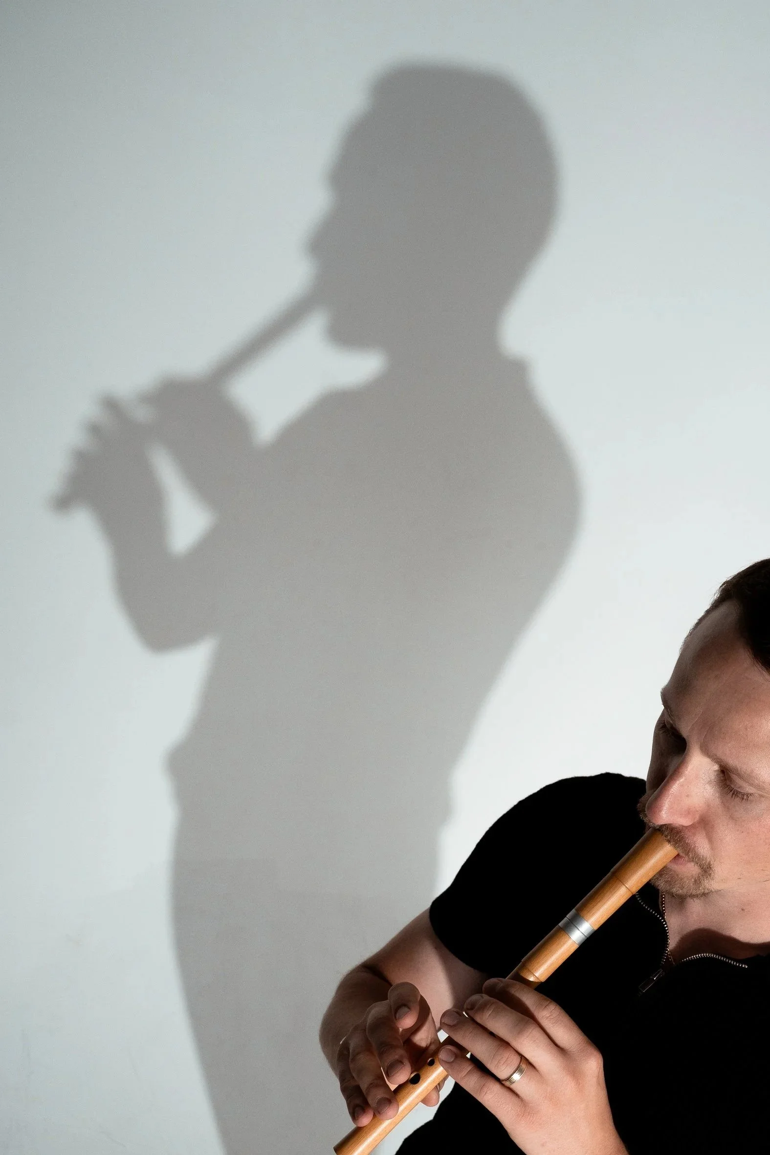 A man playing a flute with a shadow of himself holding a beer bottle projected on the wall behind him.