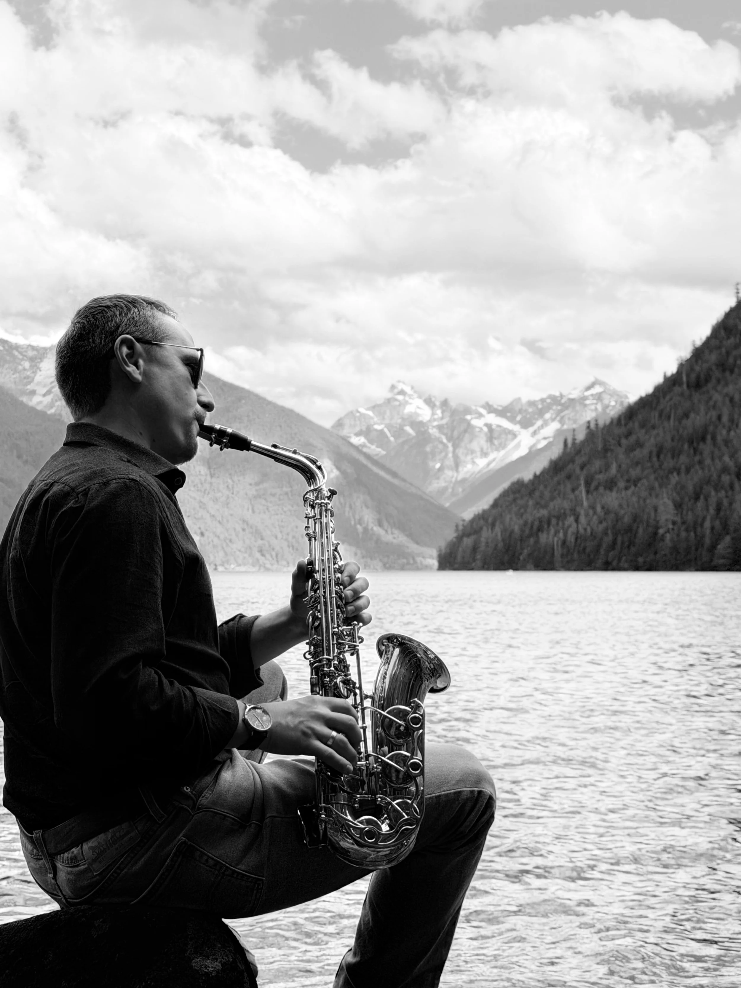 A man with short hair, wearing sunglasses and a long-sleeve shirt, playing a saxophone near a lake. The background features mountains with some snow and a partly cloudy sky.