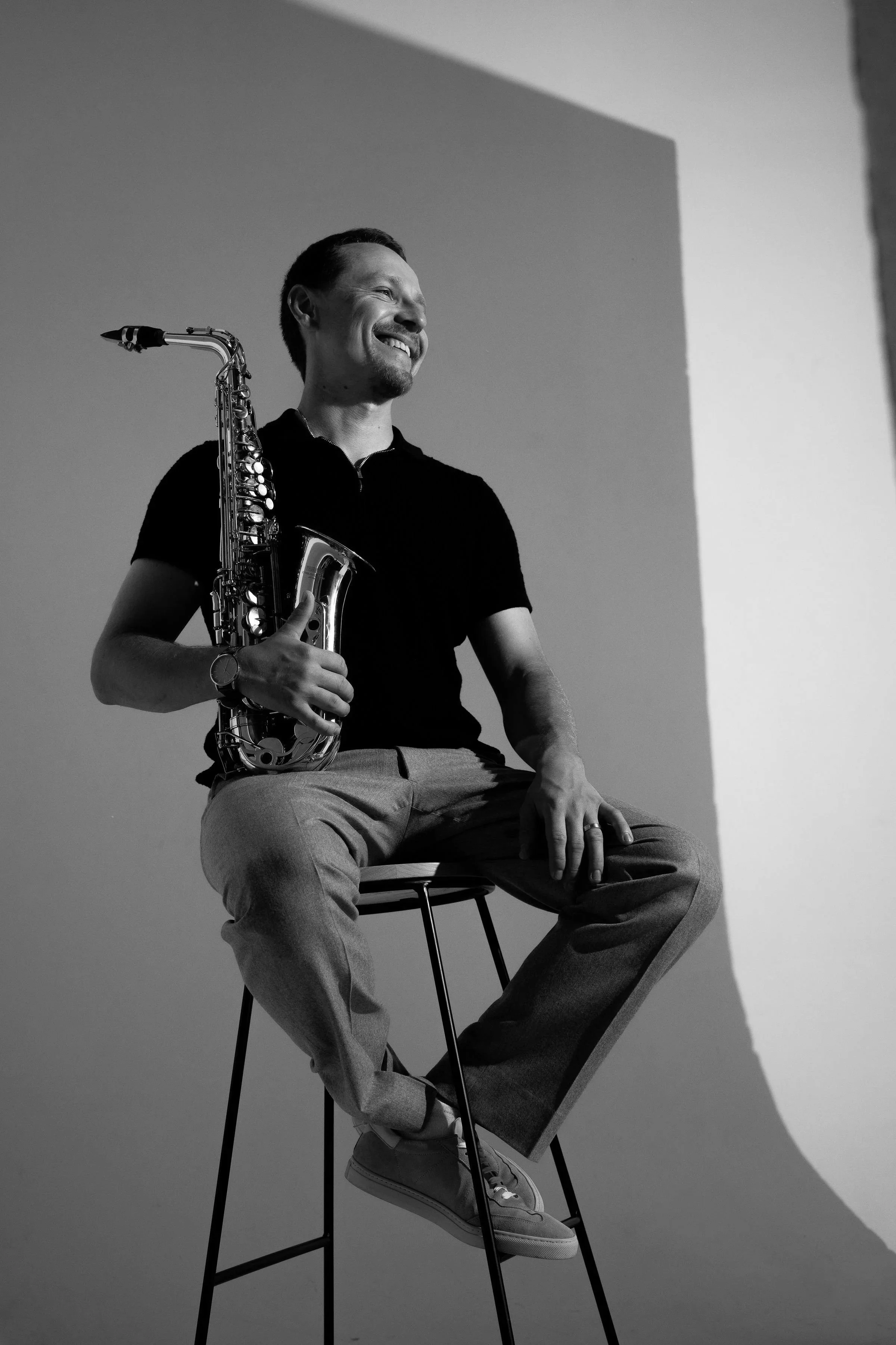 Black and white photo of a man sitting on a tall chair, holding a saxophone, smiling and looking away, with his legs crossed and wearing casual clothing.