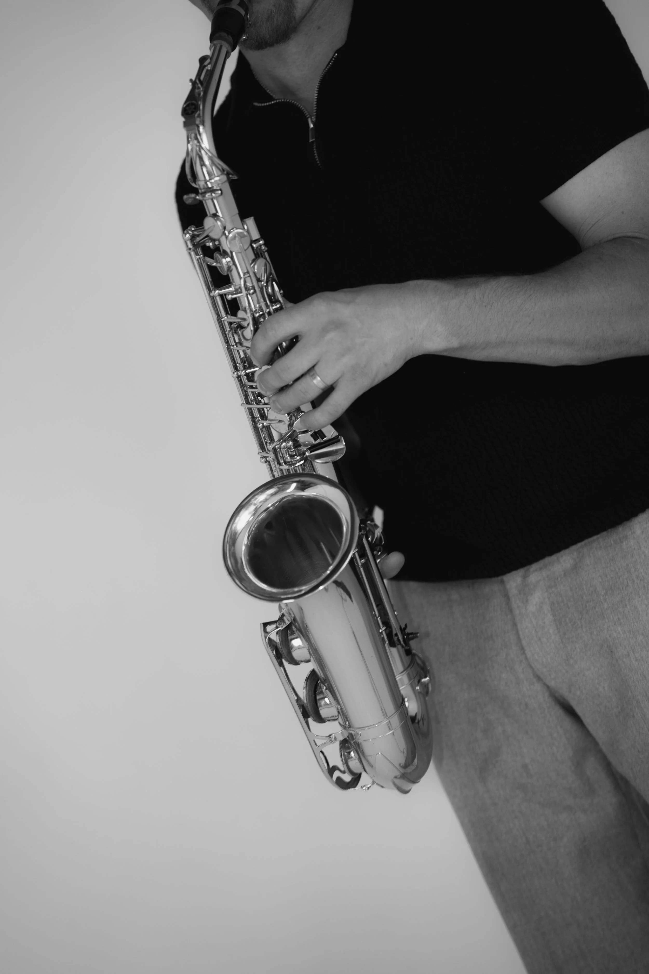 A person holding a saxophone, seen from the side, wearing a dark shirt and grey pants, with a plain light-colored background.
