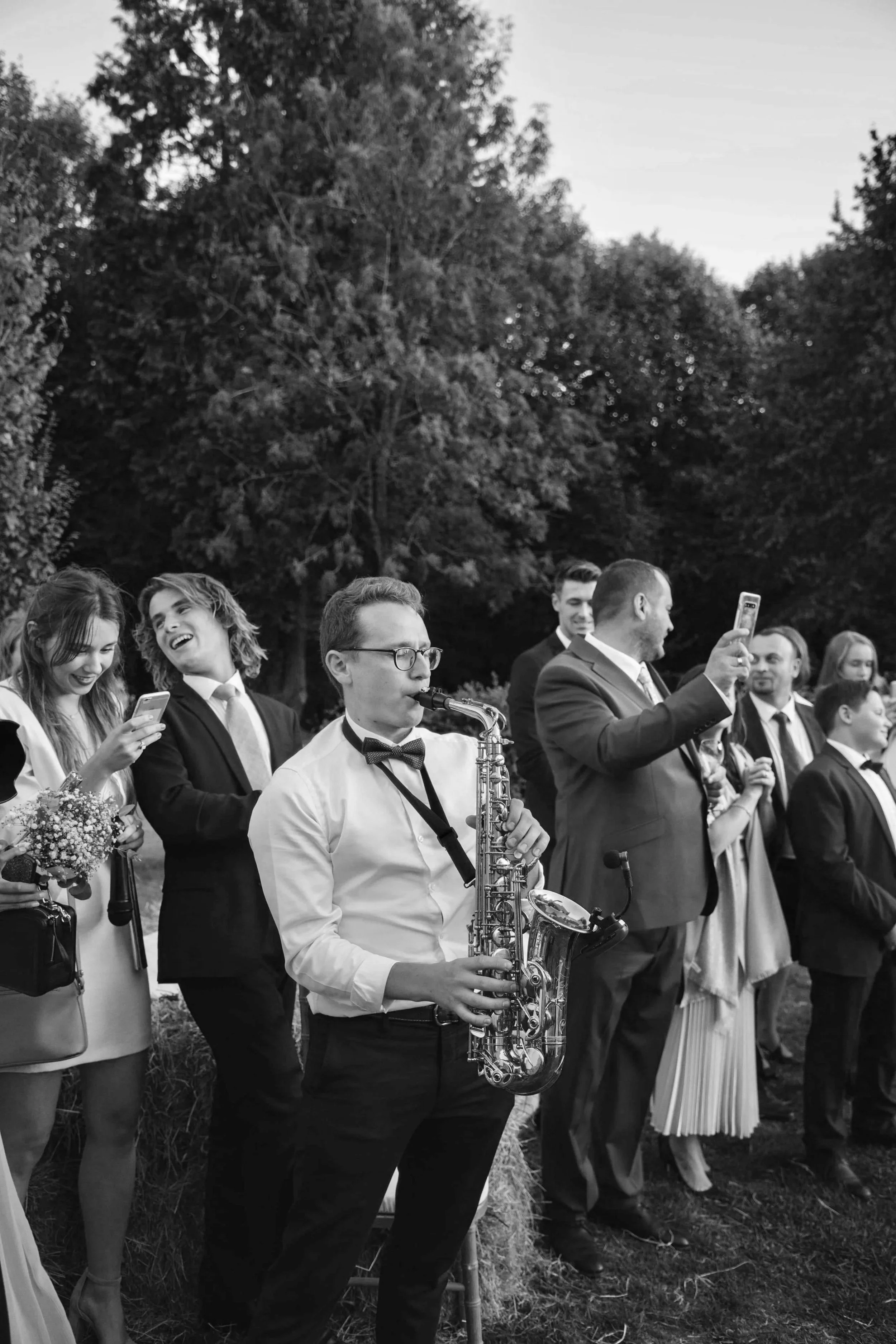 A black and white photo of a group of people at an outdoor event, with some individuals holding phones and a musician playing a saxophone.
