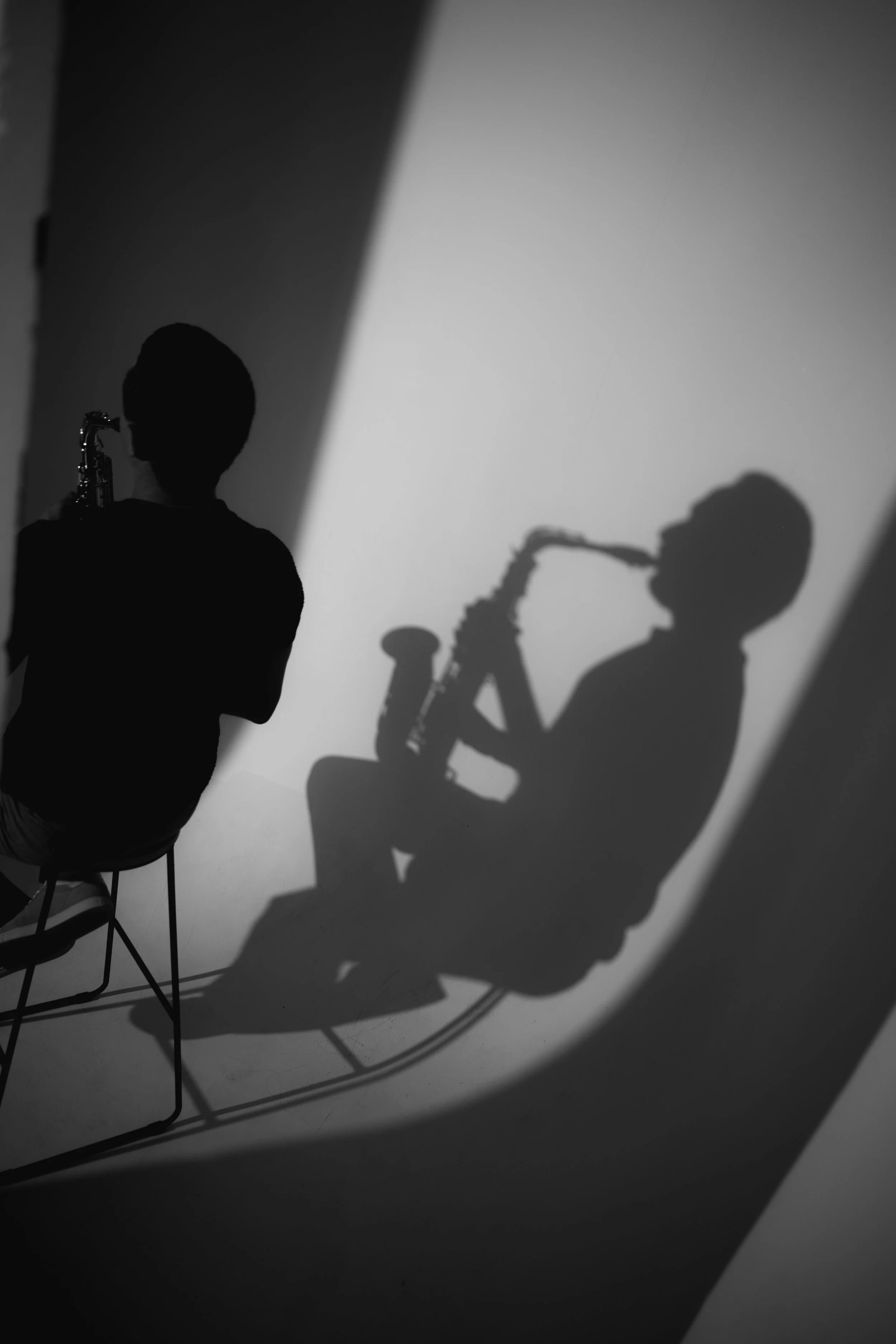 Black and white photograph of a person sitting on a chair playing the saxophone, with their shadow cast on the wall behind them.