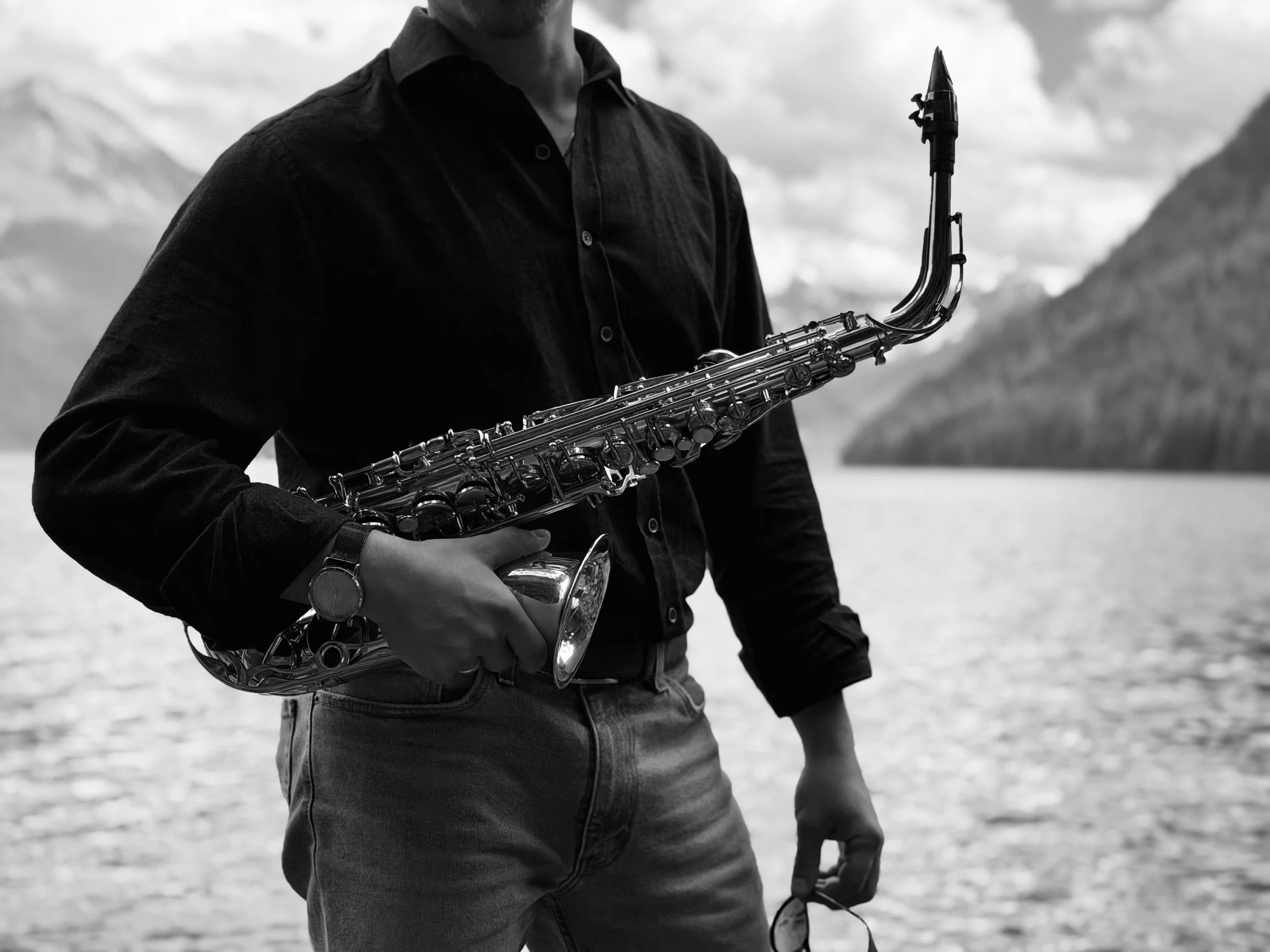 A person wearing a dark shirt and jeans, holding a saxophone, standing outdoors near a body of water with mountains in the background.