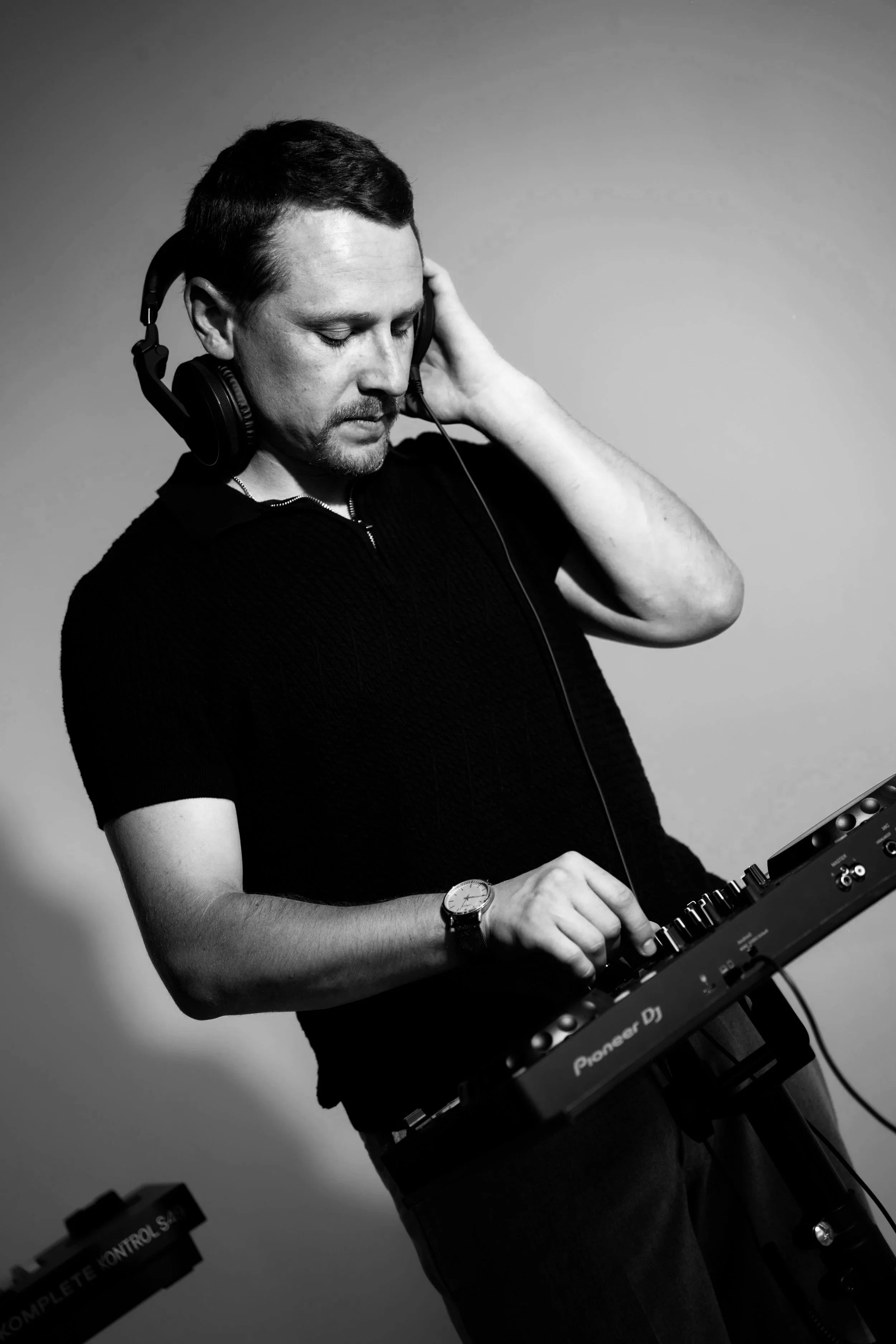 A man with short dark hair and a beard, wearing a black shirt and a wristwatch, is DJing using a Pioneer DJ controller. He is wearing large over-ear headphones and has his eyes closed, focused on his music.