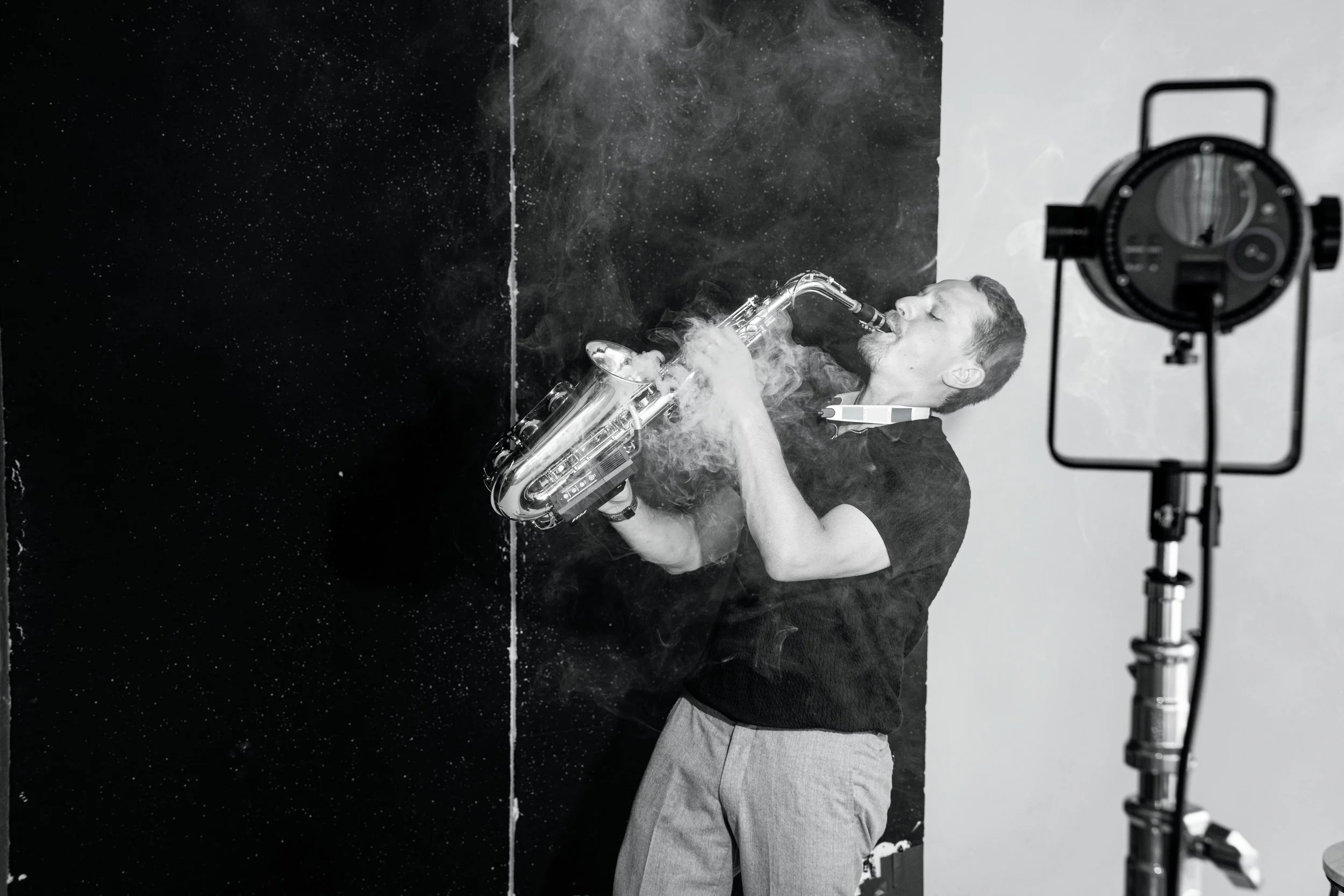 A man playing a saxophone in a photo studio with a black and white background, illuminated by a studio light on a stand.