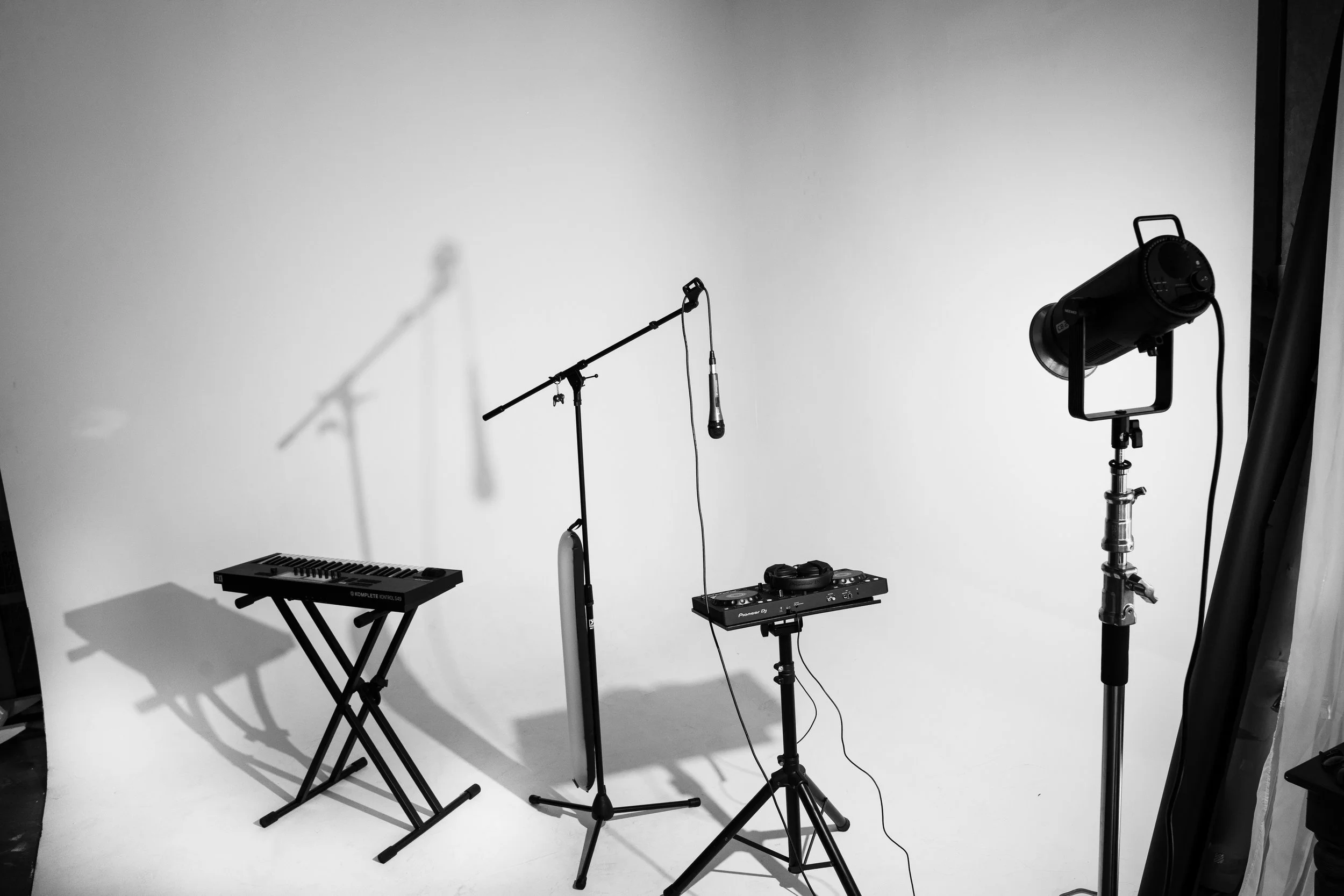 Music equipment set up in a photography studio, including a microphone on a stand, a keyboard on a stand, a DJ controller, a studio light, and a shadow of microphone on the white wall.