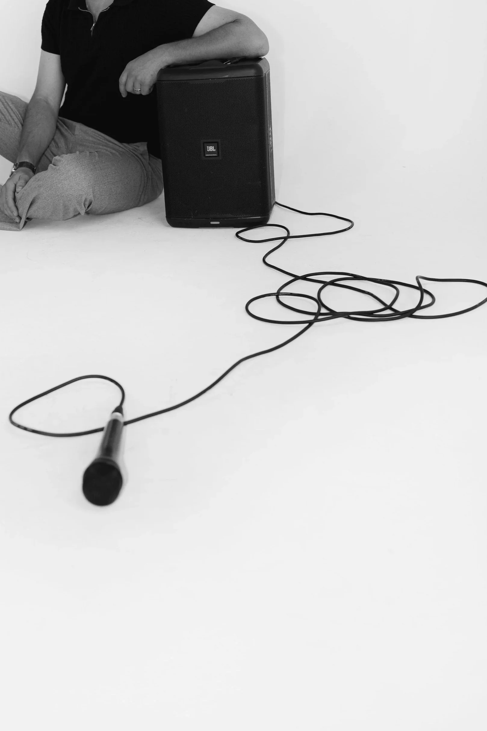 A person sitting on the floor with a large black JBL speaker beside them, holding a microphone connected to the speaker by a long cord.