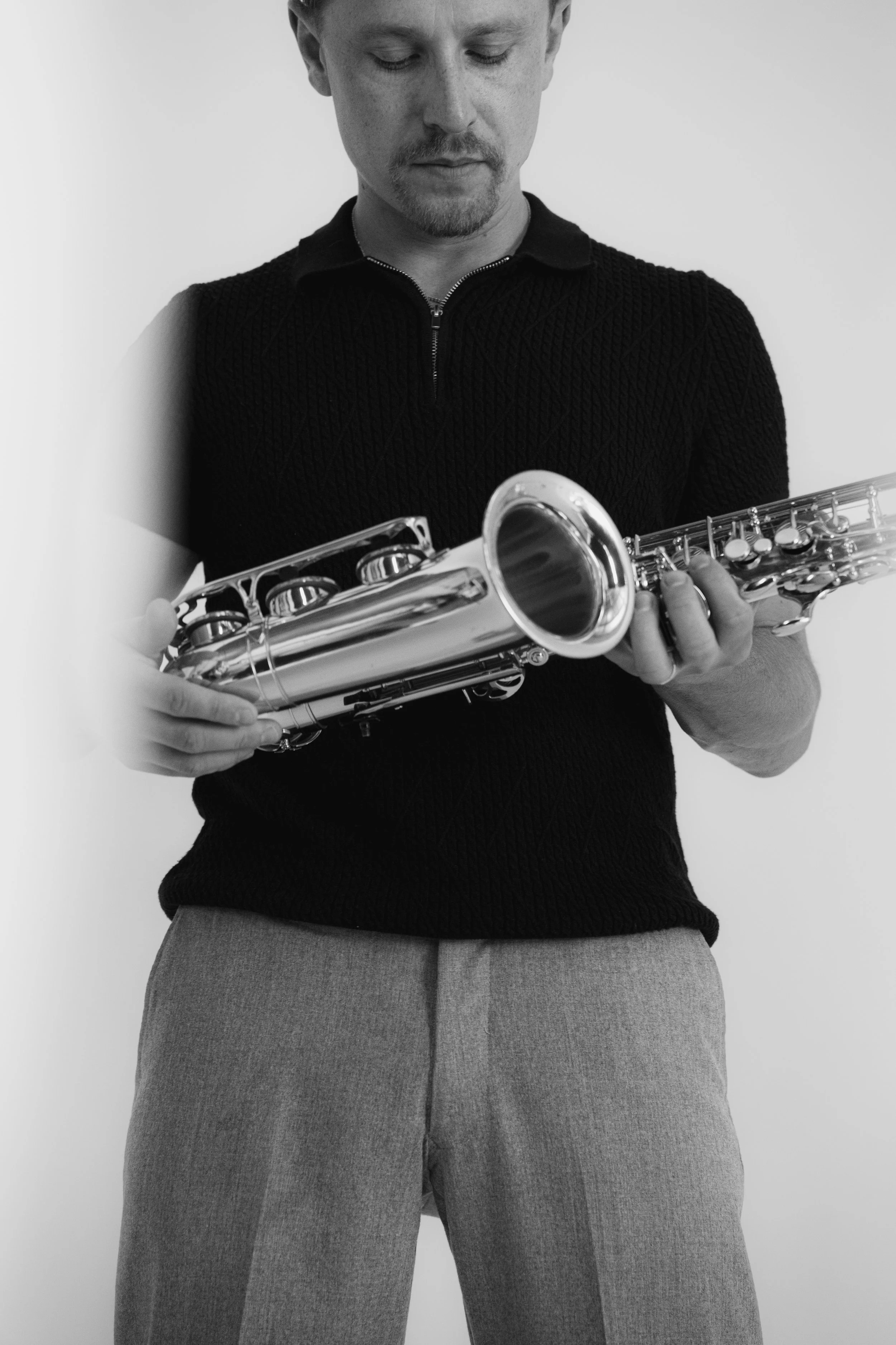 A man wearing a black textured shirt and light-colored pants holding a silver saxophone.
