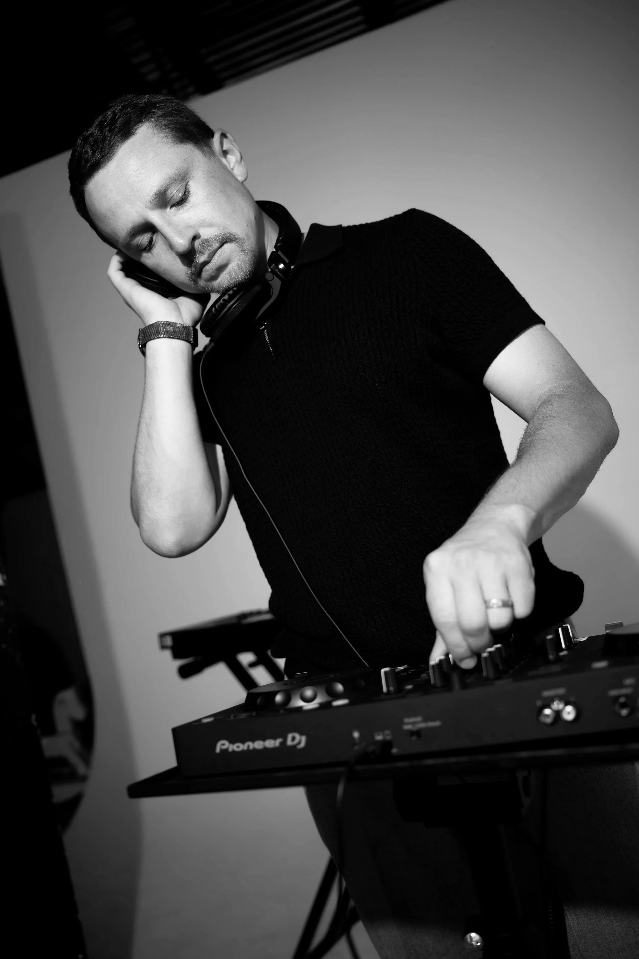A man with short dark hair, wearing a black shirt, is DJing using a Pioneer DJ controller. He is wearing headphones around his neck and appears to be adjusting the equipment while focusing on his work.