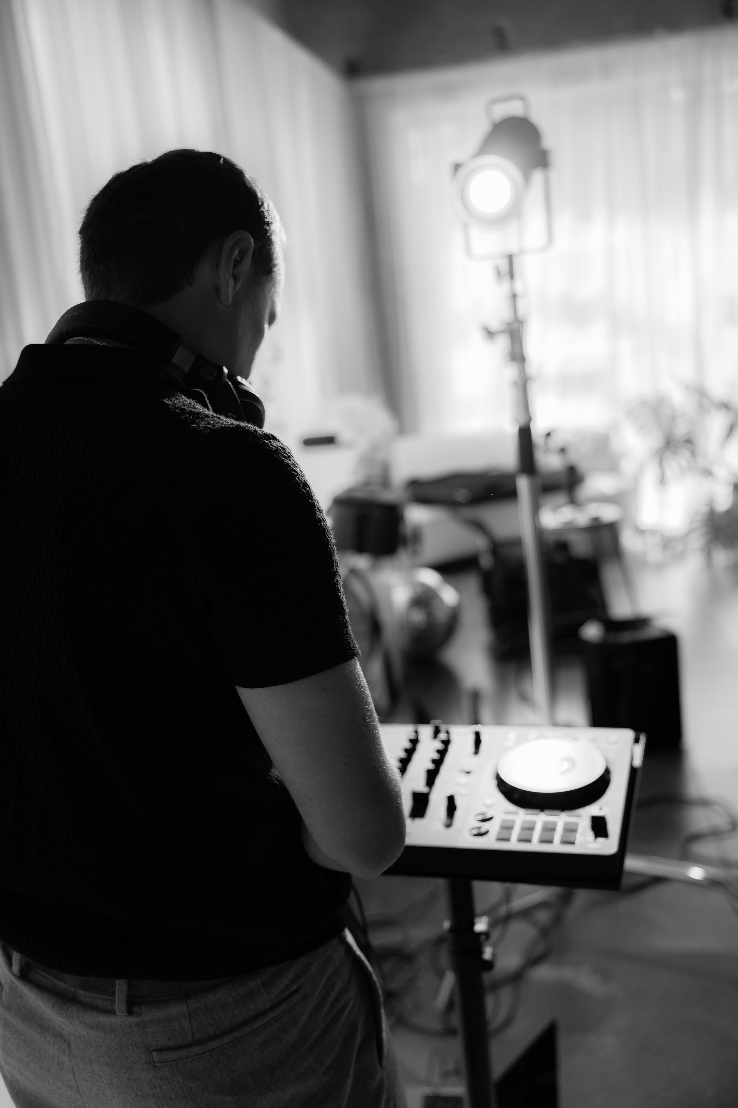 A person with dark hair wearing a black shirt and gray pants is operating a DJ mixer and turntable setup indoors, with studio lighting in the background.