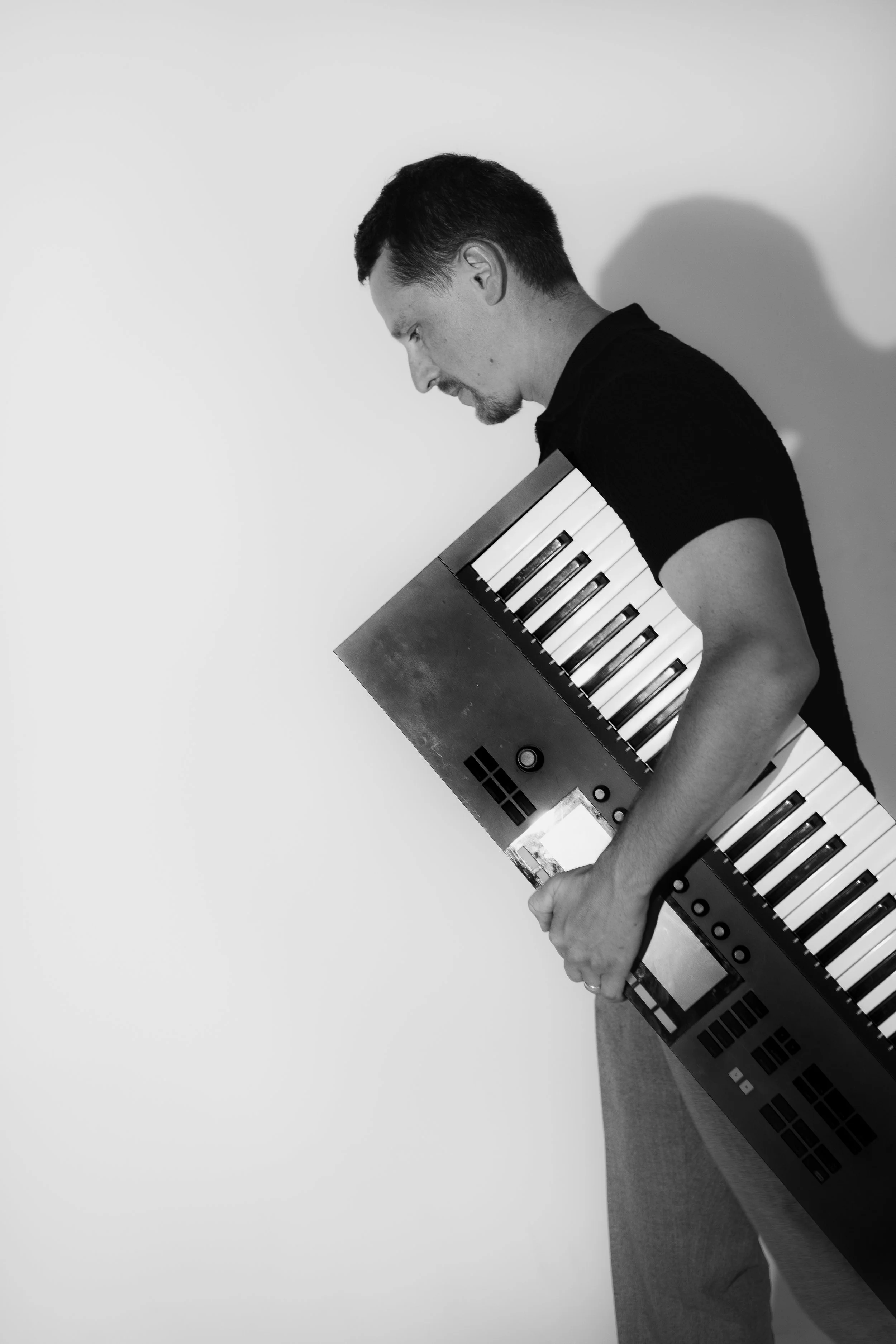 A man with a dark shirt and short hair holding a keyboard vertically against his body, looking down in front of a plain wall.