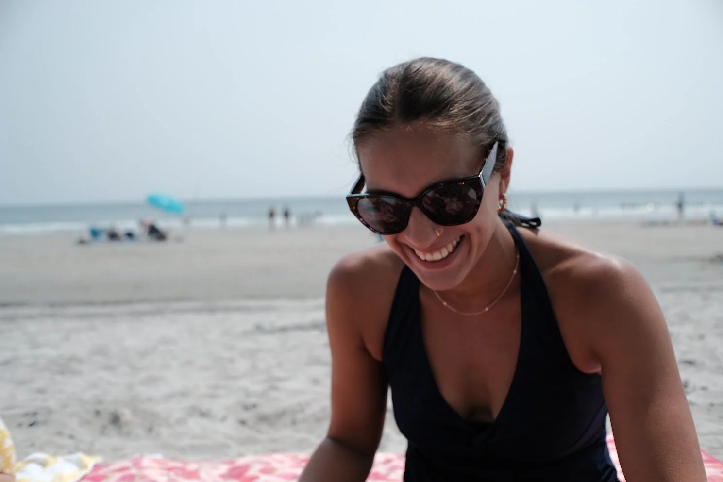 A woman wearing sunglasses and a black swimsuit smiling on a beach, sitting on a pink and yellow towel with the ocean and beach umbrellas in the background.