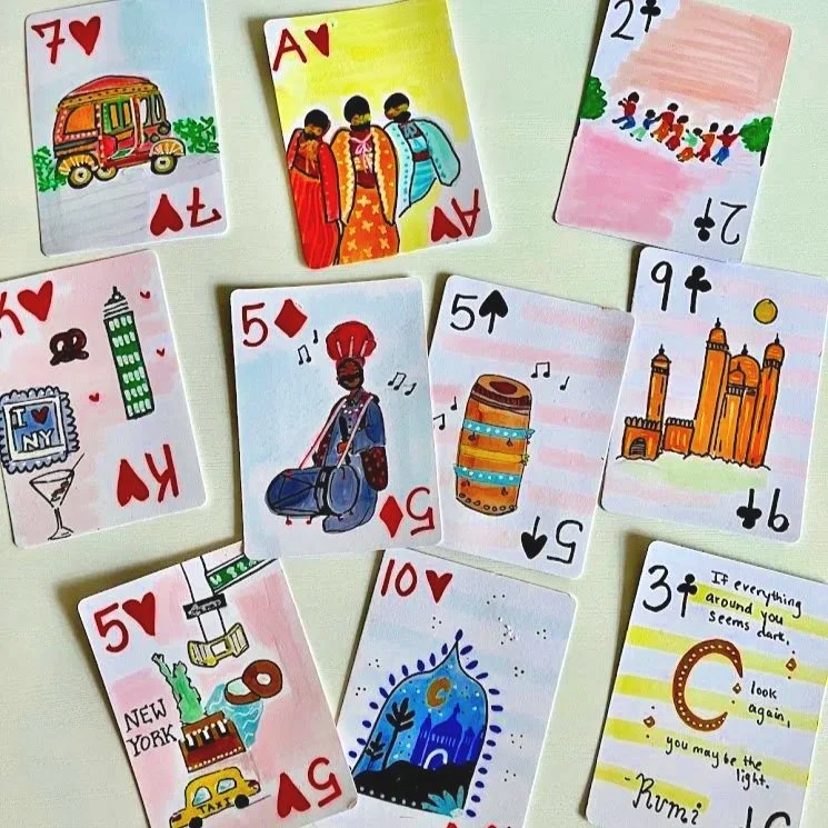 A collection of colorful playing cards laid out on a white surface, including cards with illustrations of a tuk-tuk, three women conversing, children playing, a tattoo, a castle, the New York City skyline, musical instruments, landmarks, and a quote about looking again.