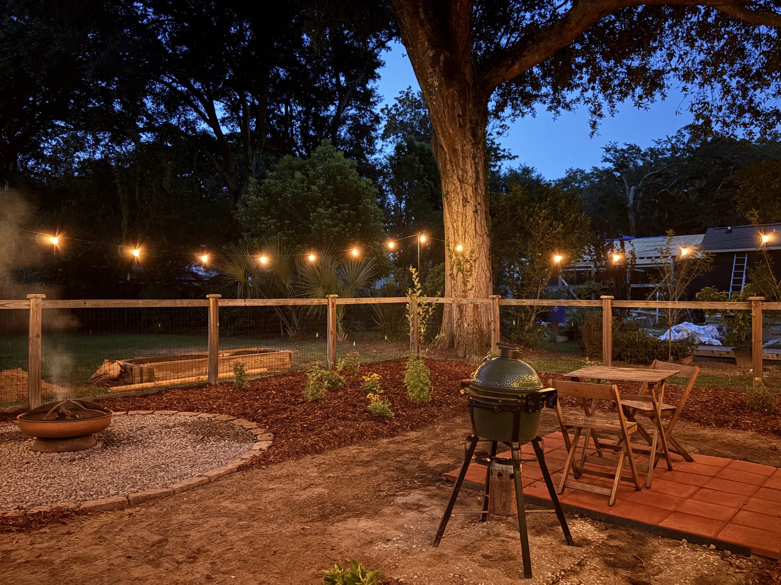 A fenced backyard at dusk featuring a firepit, grill, and dining patio with string lights hanging above. Live oak in the background along with a native plant garden.