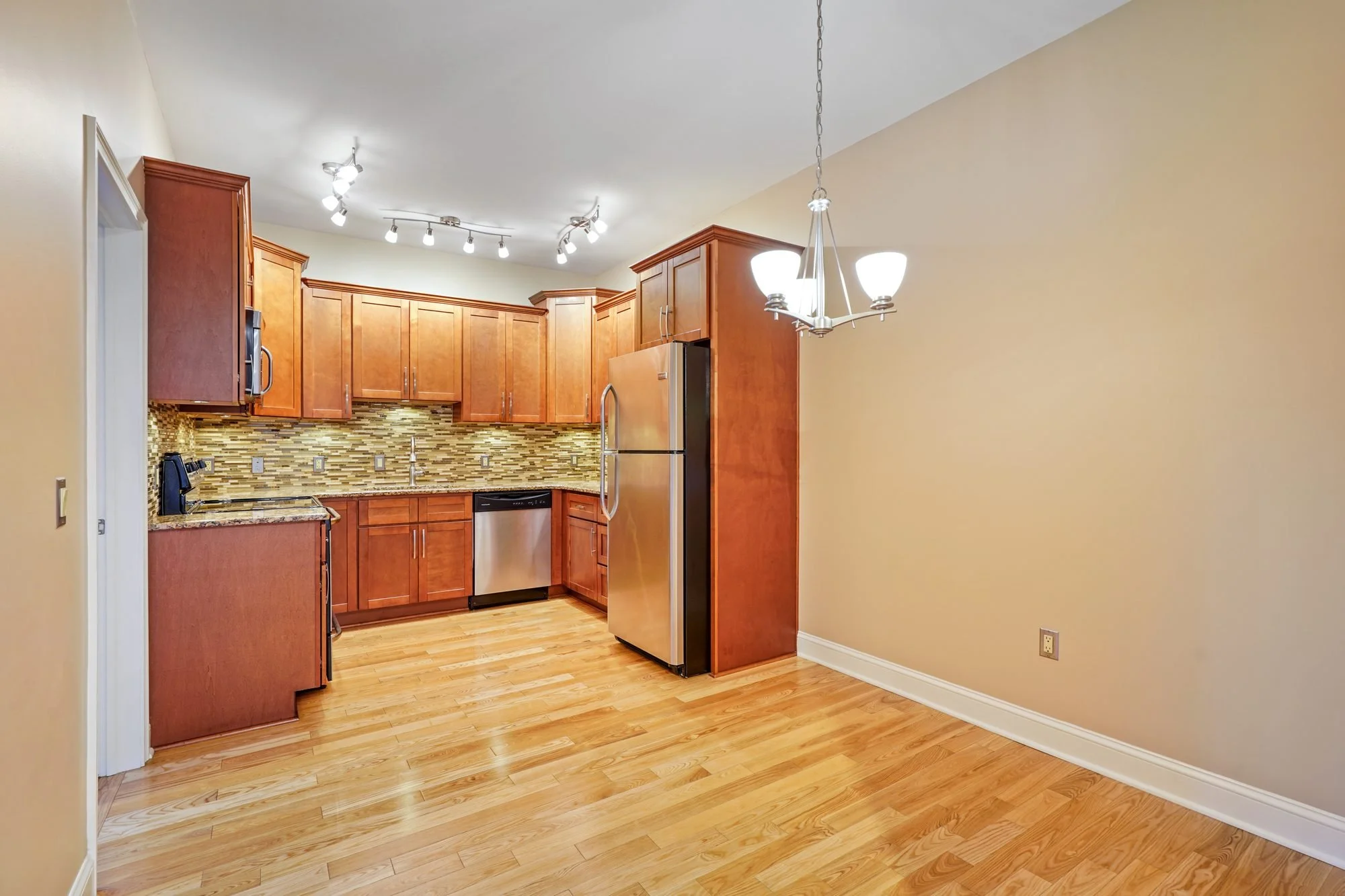 A kitchen with wooden cabinets, a stainless steel refrigerator, a mosaic tile backsplash, and a wooden floor, viewed from an adjacent room with beige walls and a chandelier hanging from the ceiling.