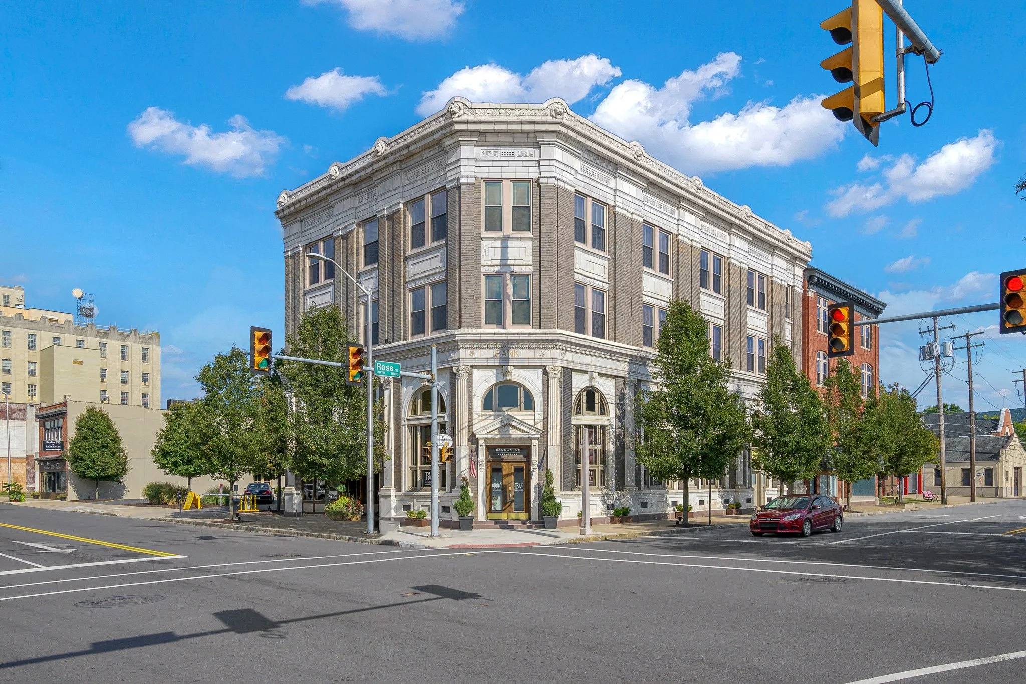 A multi-story ornate building on a city street corner with a retail store on the ground floor, traffic lights, and trees along the sidewalk under a partly cloudy sky.