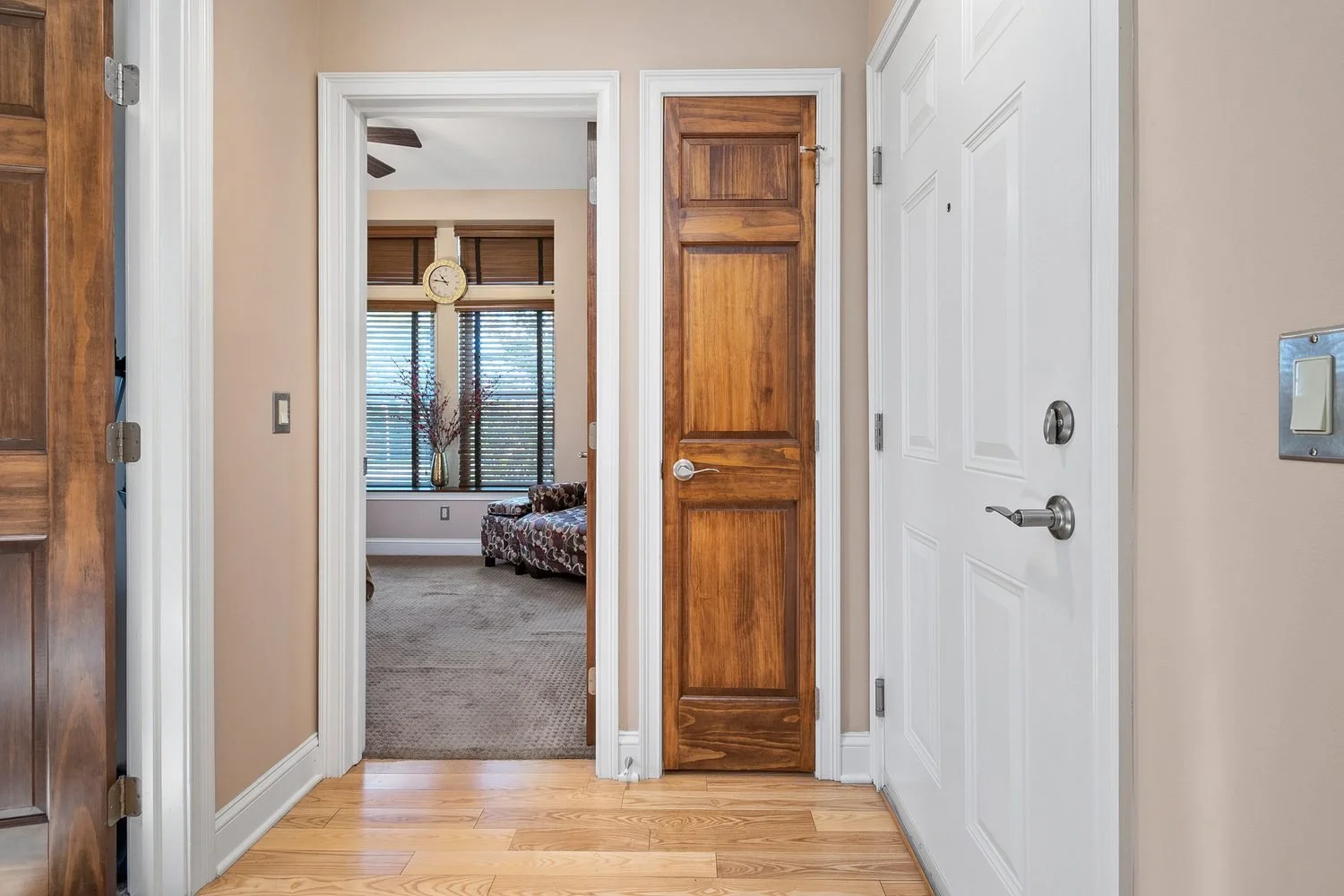 View from an entryway into a living room, showing a beige wall, a white door, a wooden closet door, and another white door. The living room has a large window with blinds, a clock, a vase with decorative branches, and upholstered chairs.